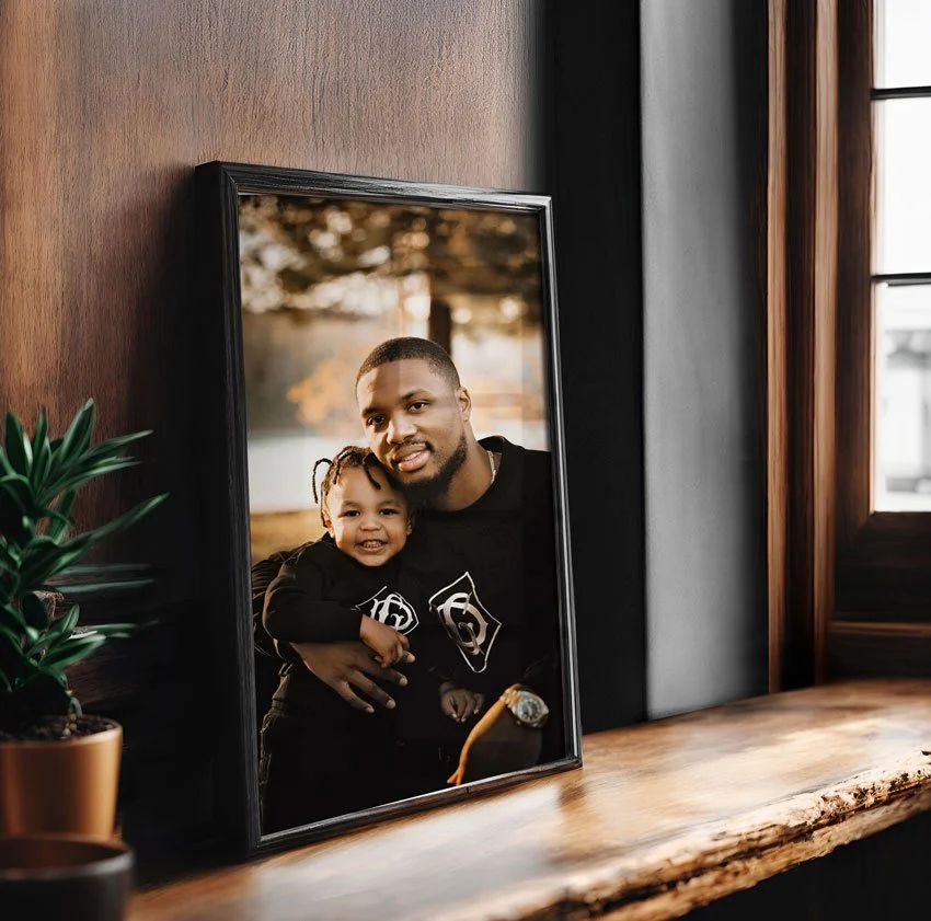 A large, black-framed professional portrait of Damian Lillard holding Dame Jr, leaning against a dark wood wall on a rustic wooden shelf.