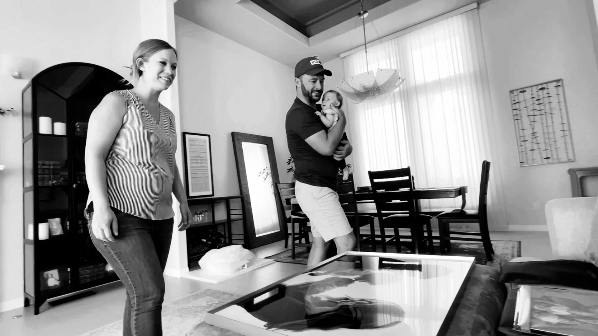 A black and white candid photo of a mother smiling and a father holding their baby while viewing a large, custom-framed portrait laid out on an ottoman during a client artwork reveal session.