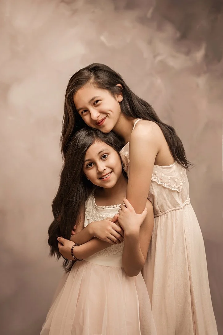 A studio portrait of two sisters with long dark hair, wearing coordinating cream lace dresses and hugging, against a warm, neutral painted background.