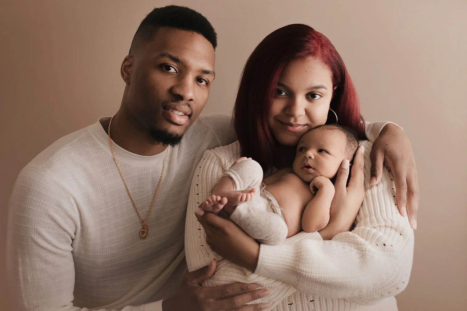 A warm and elegant studio newborn family portrait by SunnyMelon Photography featuring NBA star Damian Lillard and his family with their infant son, Dame Jr. The image shows the family dressed in clean, neutral white tones against a soft, earth-toned 