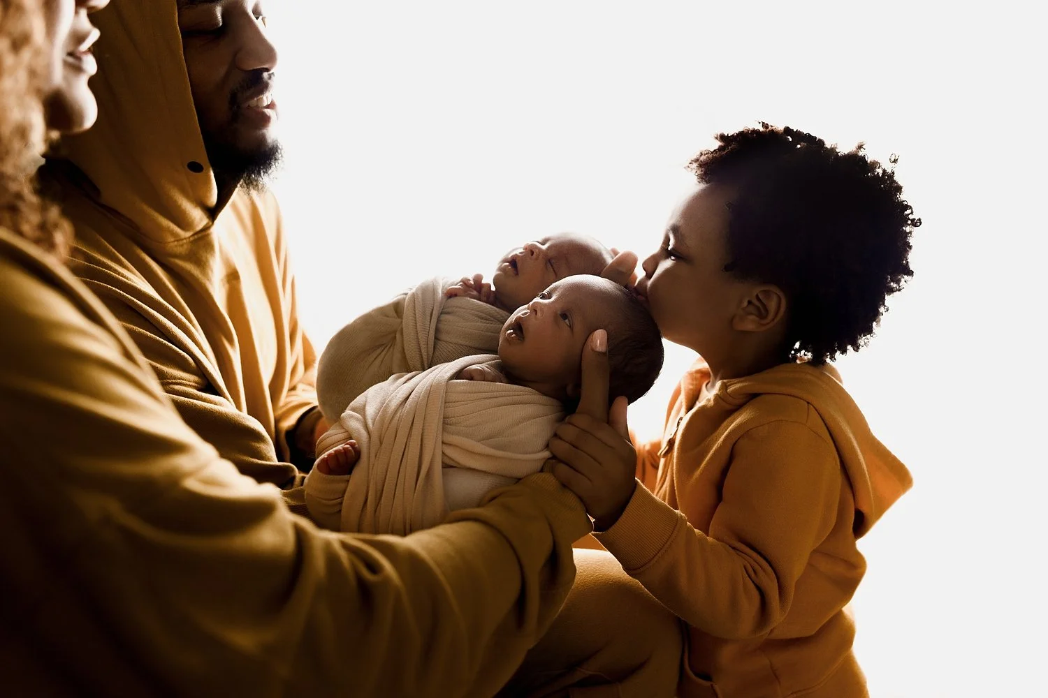 A high-contrast, backlit silhouette-style image of Damian Lillard holding his two newborn twins, who are wrapped in cream swaddles.

Big brother Dame Jr. is shown tenderly kissing the forehead of one of the twins.

The family is coordinated in warm, 