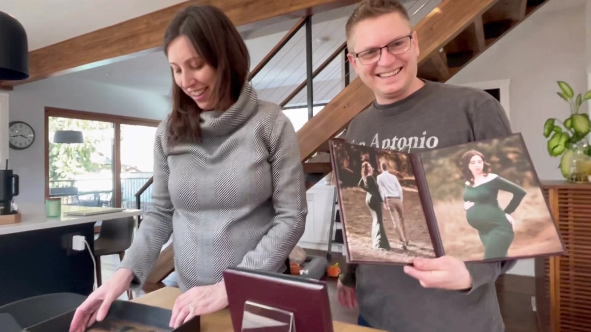A happy man and pregnant woman in their home, smiling while holding open a professional maternity photo album featuring outdoor portraits.