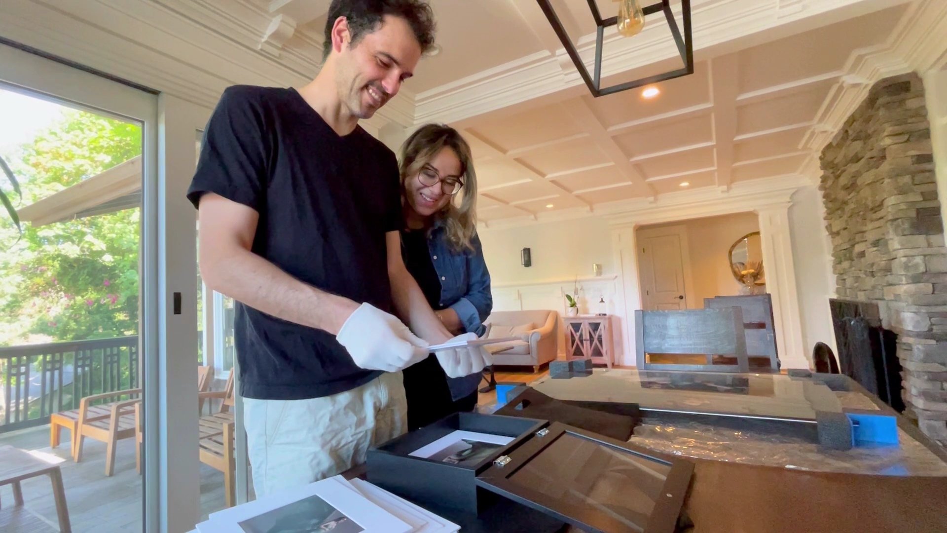 A smiling couple in a bright, modern living room views high-quality matted prints from a luxury black presentation box. The father wears white handling gloves while looking at the photos, showcasing the premium final products and boutique reveal serv