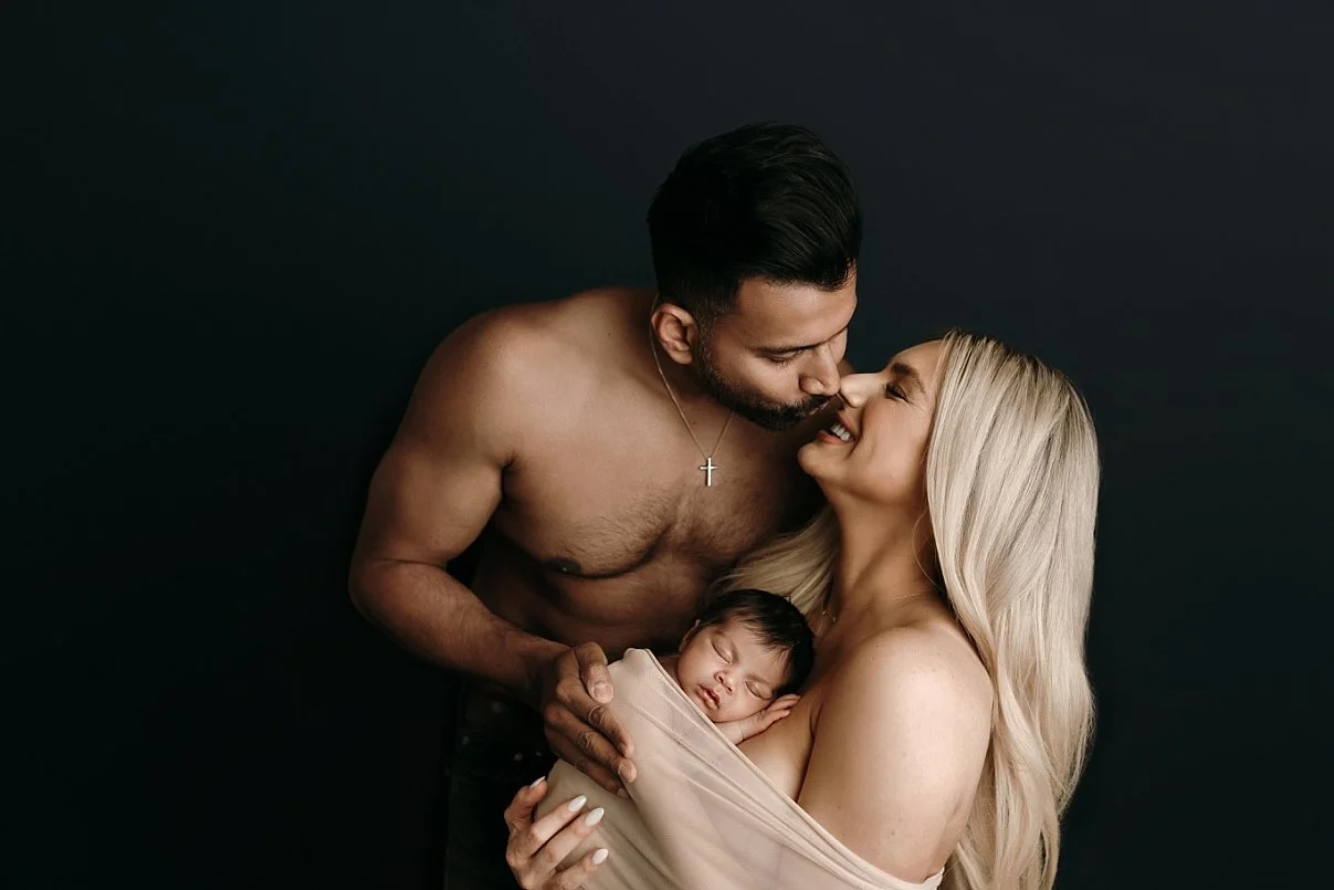 An intimate family photograph featuring a shirtless father with a gold cross necklace leaning in to kiss the mother. The mother, with long blonde hair, smiles up at him while holding their infant, who is peacefully asleep and swaddled in a neutral ta