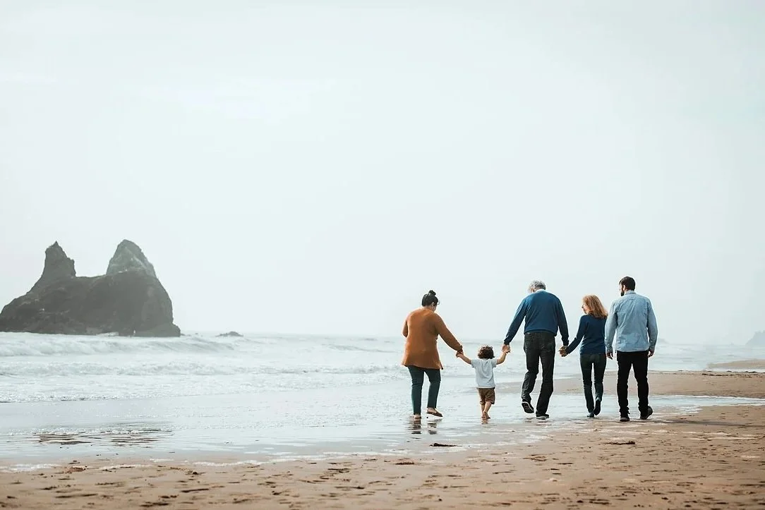 A family of five, including grandparents and a small child, walk hand-in-hand along the shoreline of a misty Oregon beach with large rock formations in the background.