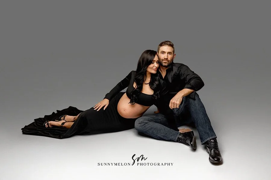 A pregnant woman in a sleek black long-sleeve crop top and matching floor-length skirt reclining next to a man in a black button-down shirt and jeans, posing against a clean grey studio backdrop.