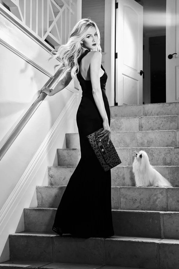 A blonde woman in a black evening gown holding a leopard print clutch poses on a stone staircase with a white Maltese dog in a high-contrast black and white editorial photo.