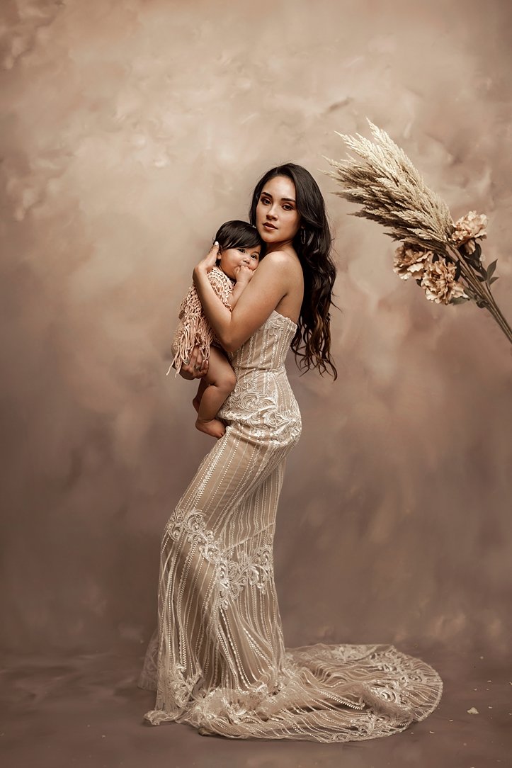 This sophisticated fine art motherhood portrait captures a mother and her young daughter in a timeless, editorial style. The mother is dressed in a stunning, intricately detailed boho-inspired lace gown, while the child is held close, creating a sens