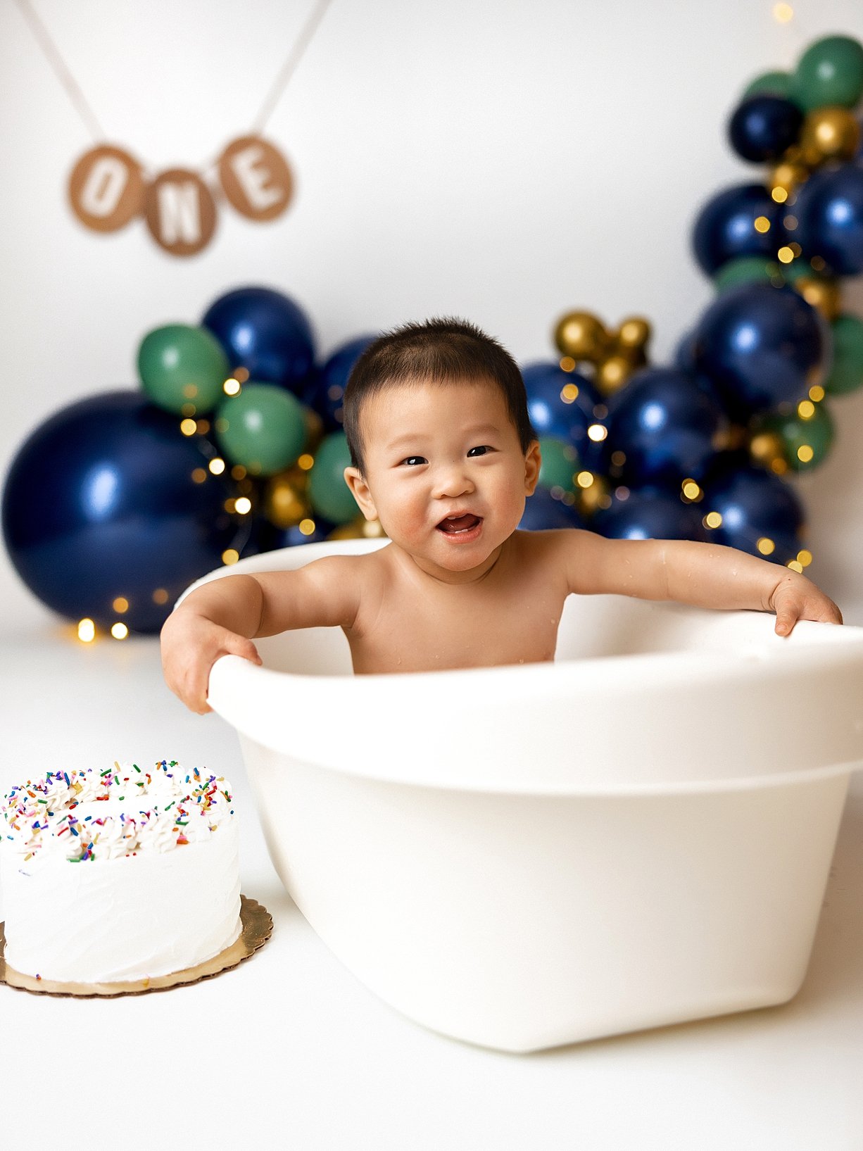 This joyful milestone portrait features the "splash" portion of a first birthday celebration, a popular way to clean up after a cake smash. The baby is captured with a happy expression inside a classic white tub, set against a sophisticated backdrop 