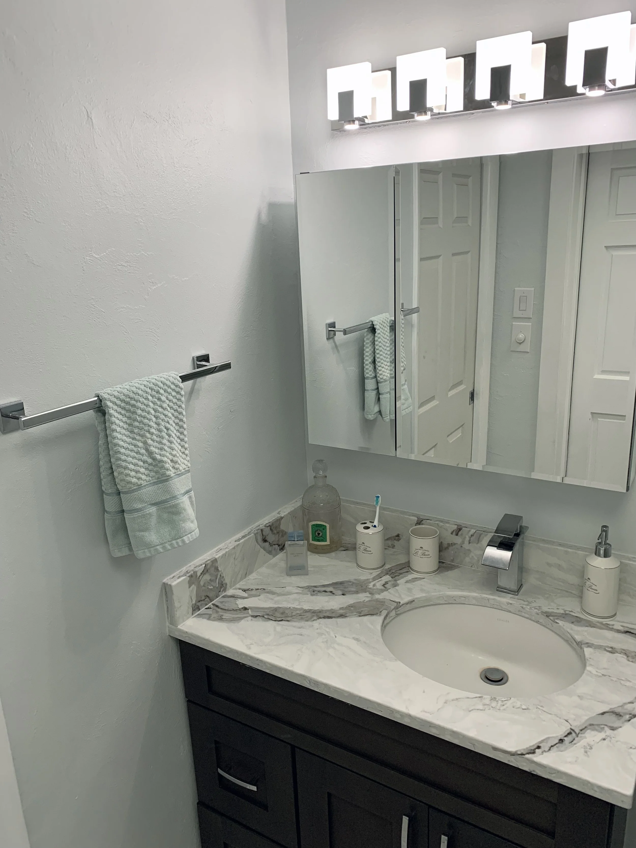 A modern bathroom vanity with a marble countertop, a round sink, and a chrome faucet. On the countertop are a glass dispenser, a toothbrush in a holder, a soap pump, and two cups. Above the vanity is a large mirror with bright wall-mounted lights. A towel is hanging on a towel bar mounted on the wall beside the mirror. The bathroom door and wall switch are visible in the reflection.