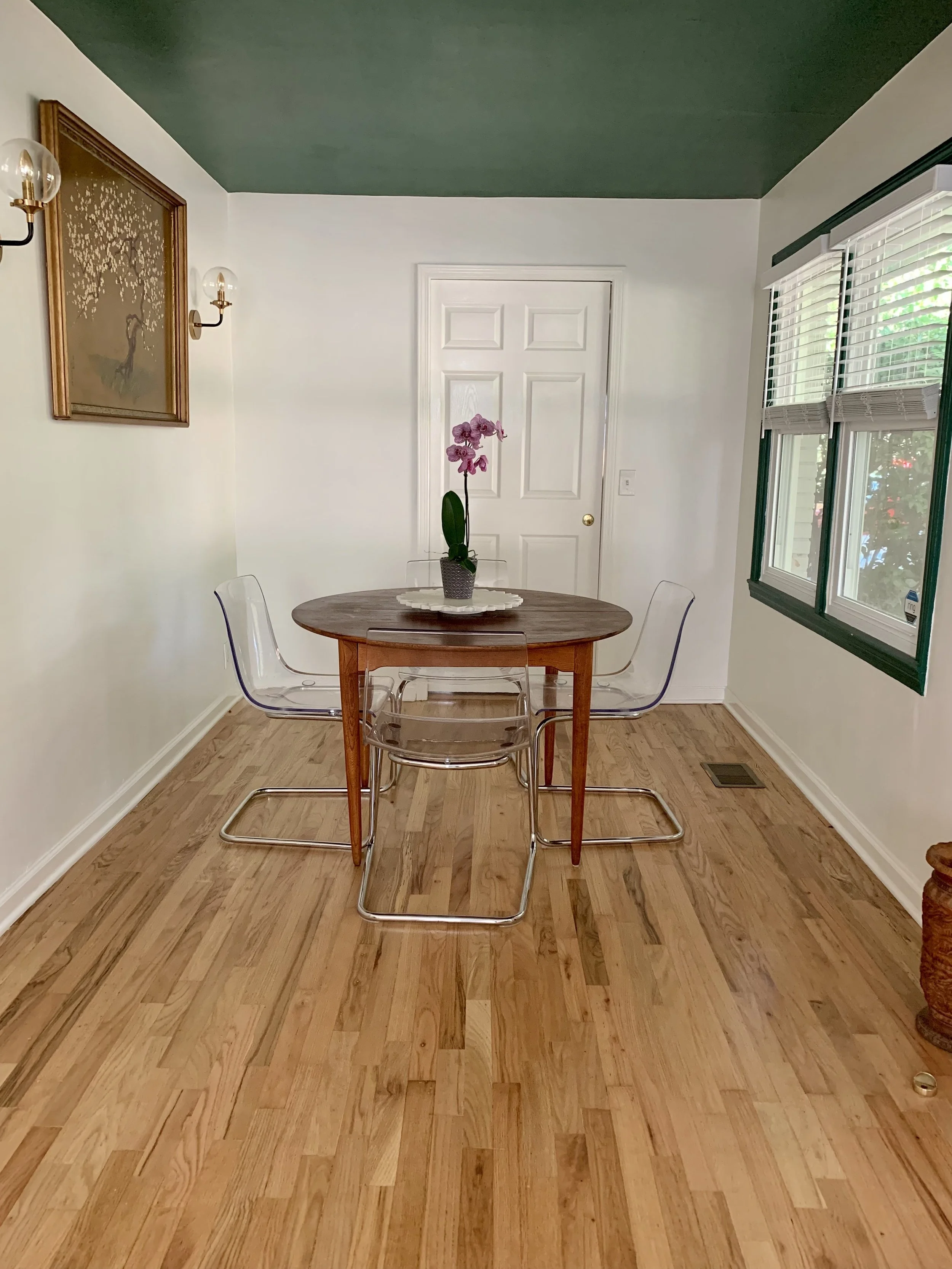 A dining room with a midcentury round wooden table, four clear acrylic chairs, a potted orchid on a white marble tray, a gold-framed antique painting of a blossoming tree on the wall, and two gold wall sconces with glass shades.

