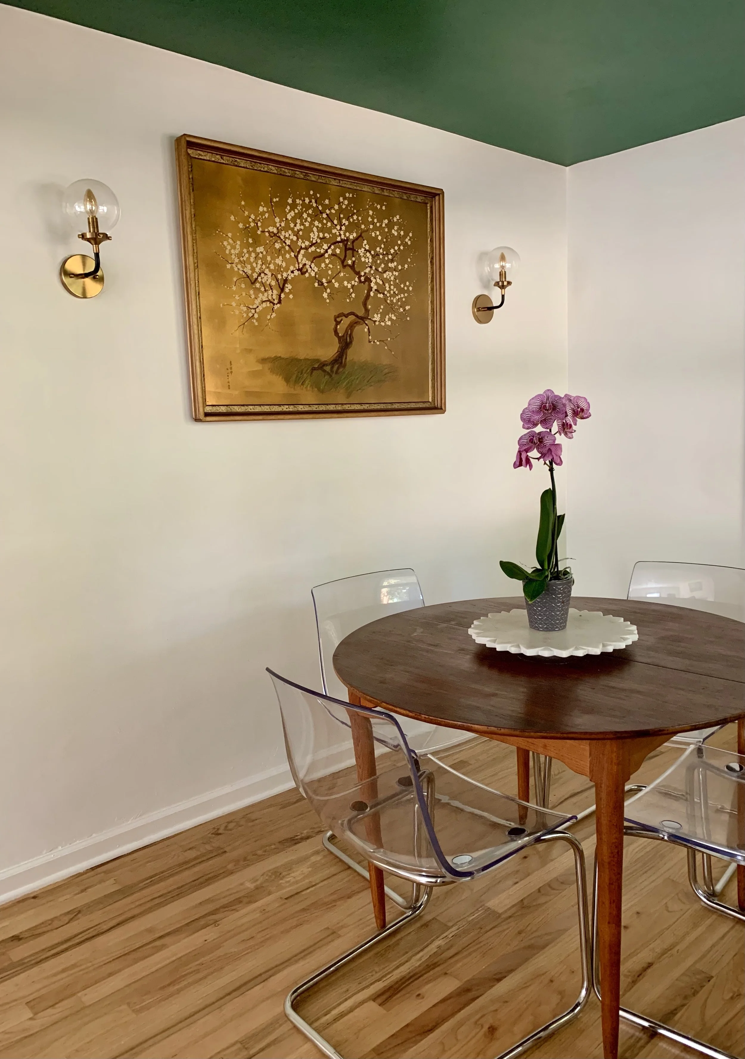 A dining room with a midcentury round wooden table, four clear acrylic chairs, a potted orchid on a white marble tray, a gold-framed antique painting of a blossoming tree on the wall, and two gold wall sconces with glass shades.