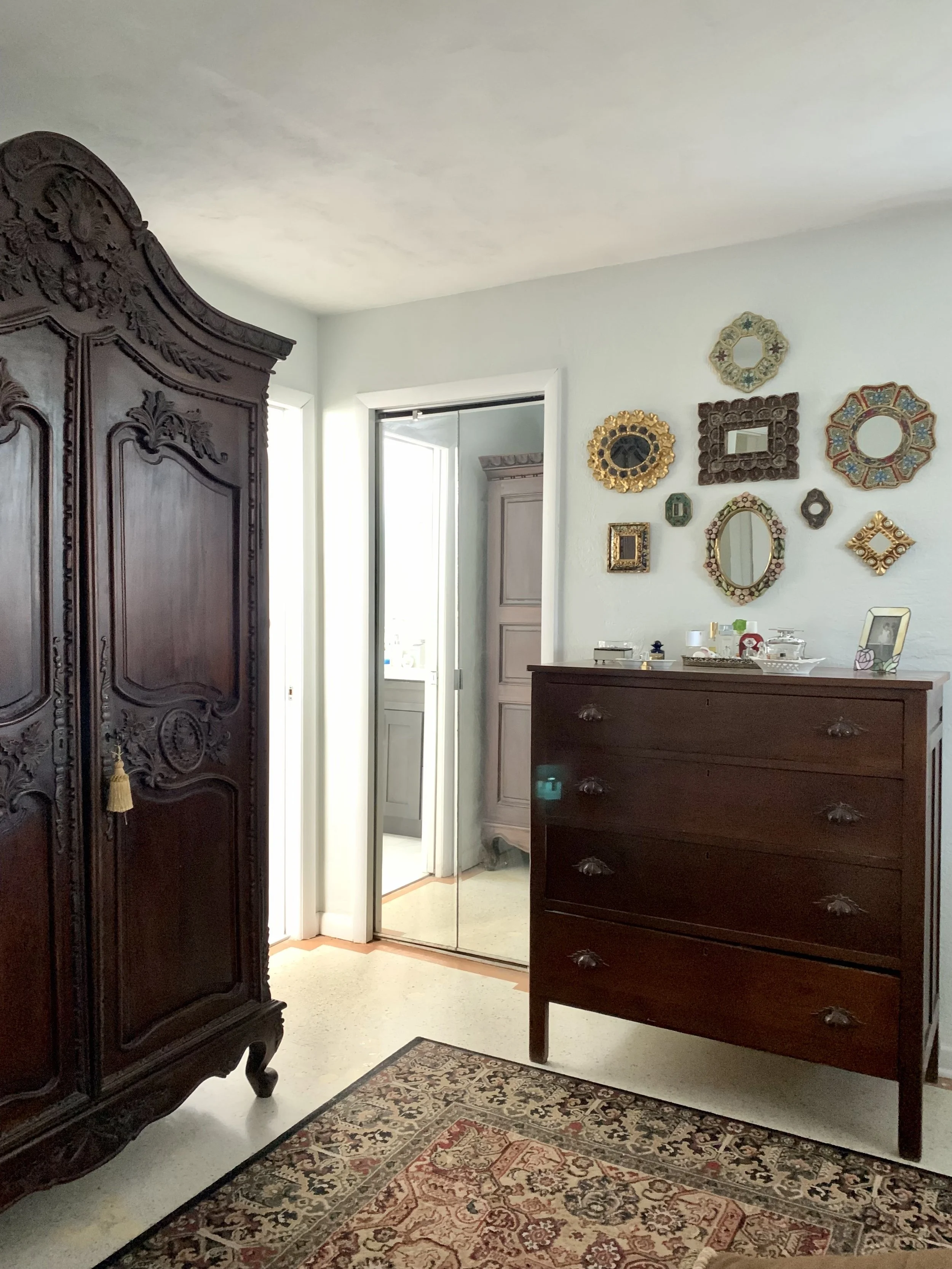 A corner of a room with an antique wooden wardrobe, a dark wooden dresser, a mirror closet with sliding doors, a window, and a wall decorated with various ornate antique mirrors and picture frames.