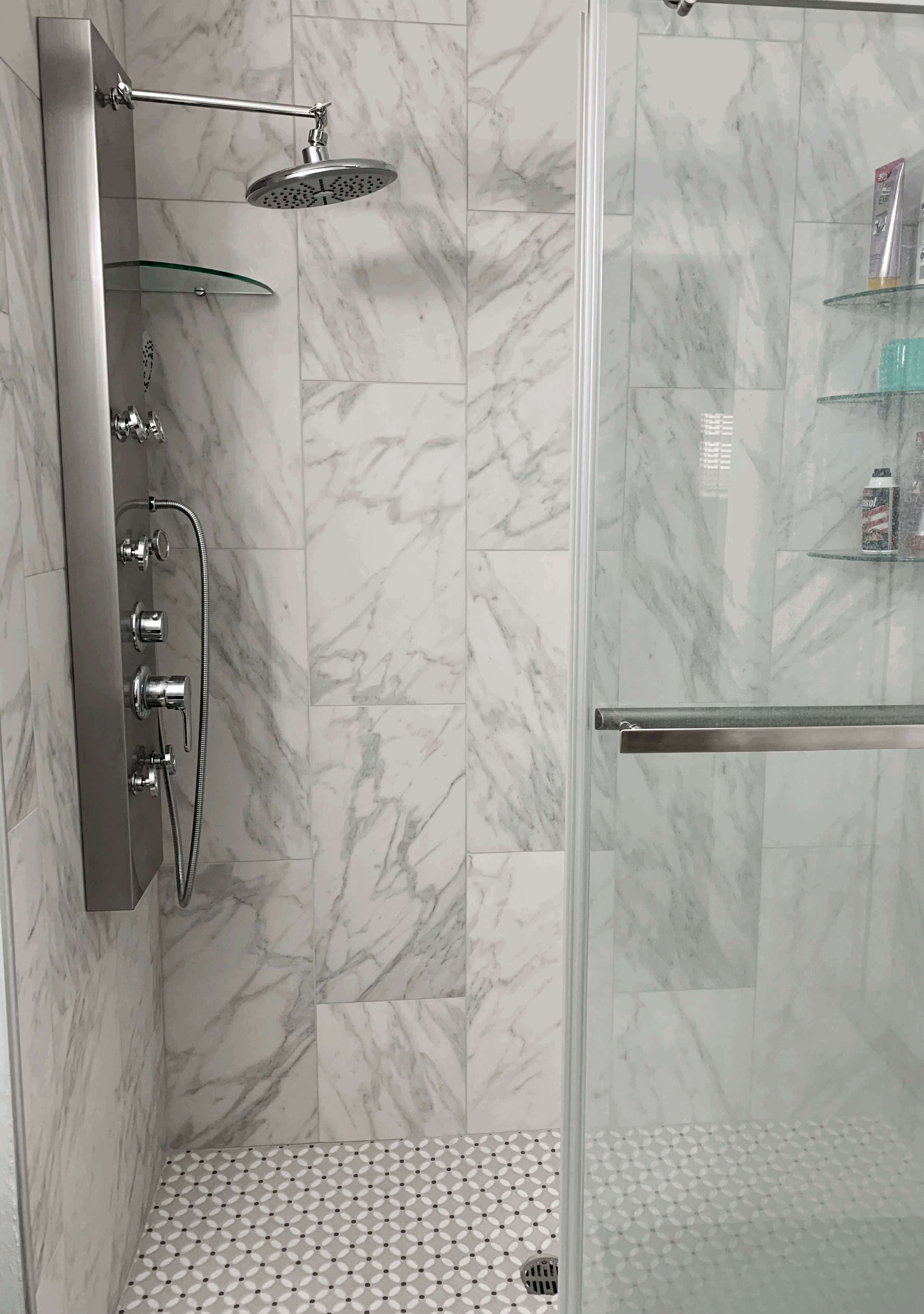 Shower with marble walls, a glass door, a wall-mounted shower panel with multiple controls, a rain shower head, and glass shelves with toiletries.