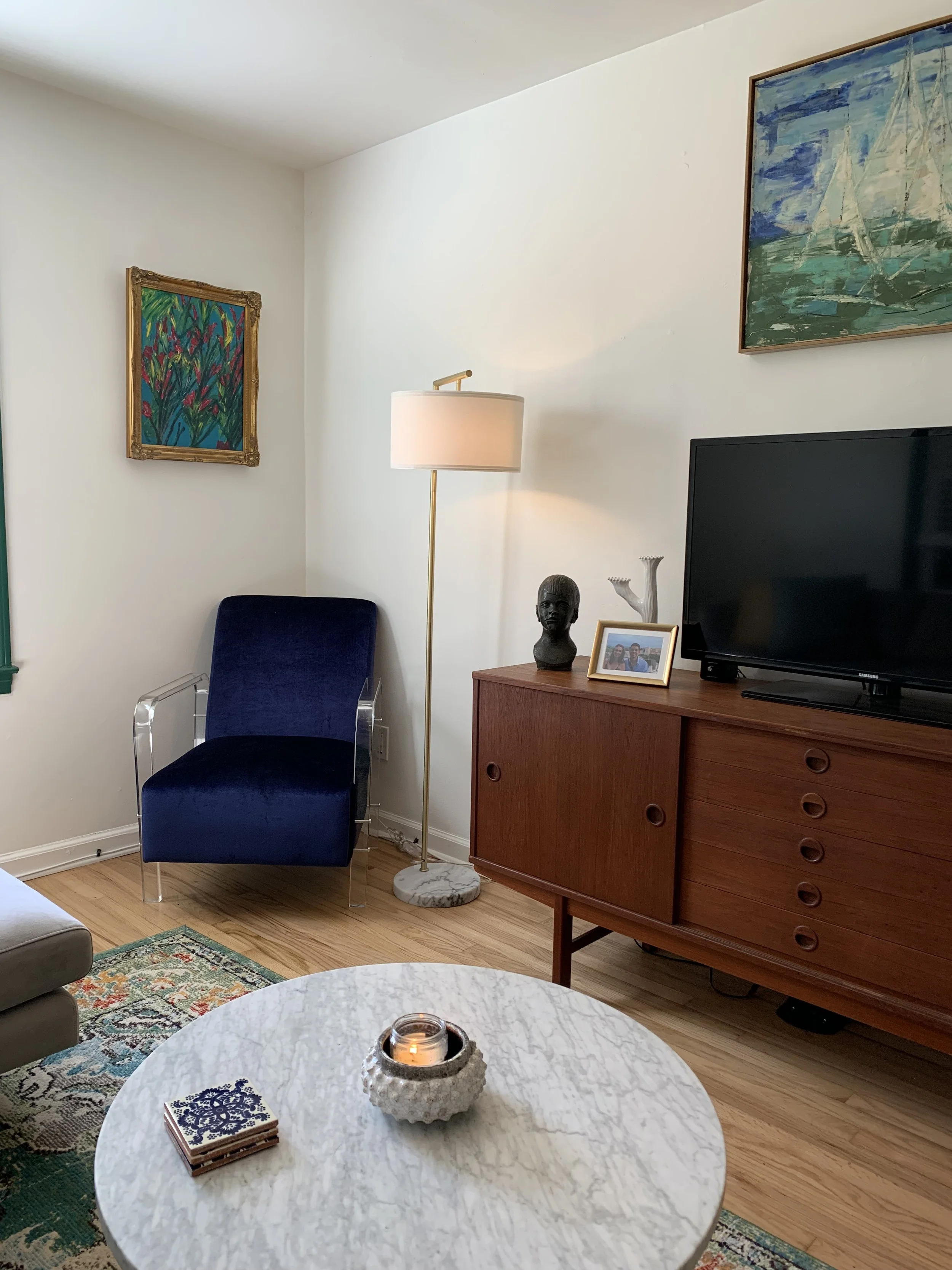 Living room corner with white walls, a blue armchair with transparent arms, a tall floor lamp with a white shade, framed paintings on the wall, a wooden cabinet with a TV, a small sculpture, a photo frame, a vase, and a marble coffee table with a candle and coasters.