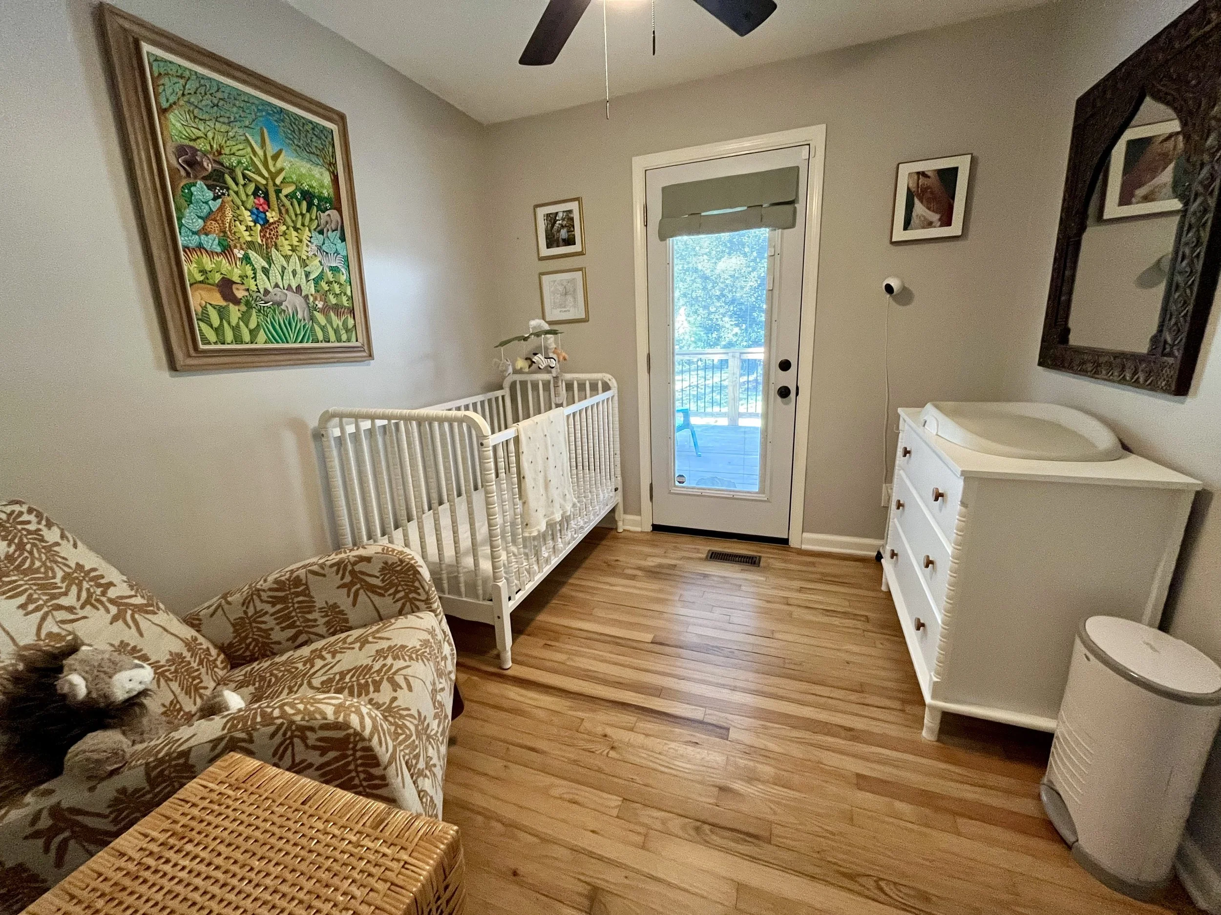 A nursery with a wooden floor, a white crib, a patterned armchair, and a white dresser with a high-changing pad. There is a door leading outside, with a view of a balcony and trees. Wall art includes a large colorful jungle scene painting and smaller framed pictures. A mirror is hung above the dresser, and a ceiling fan is visible.