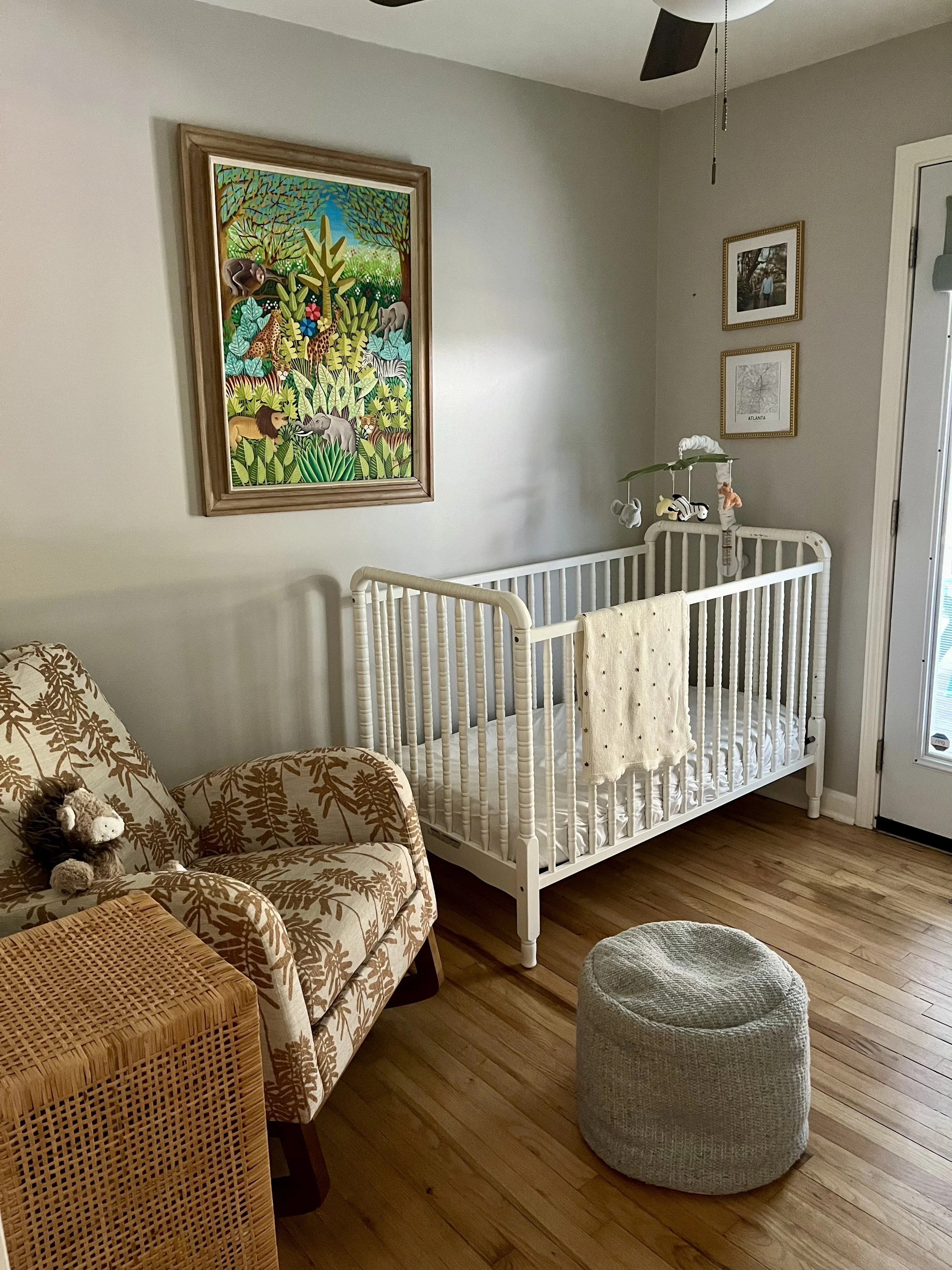 A nursery room with a white crib, a beige upholstered armchair with a animal plush toy, a round knitted ottoman, and wall art including a colorful jungle scene and a framed black and white photo. There's a ceiling fan and a glass door to the right.