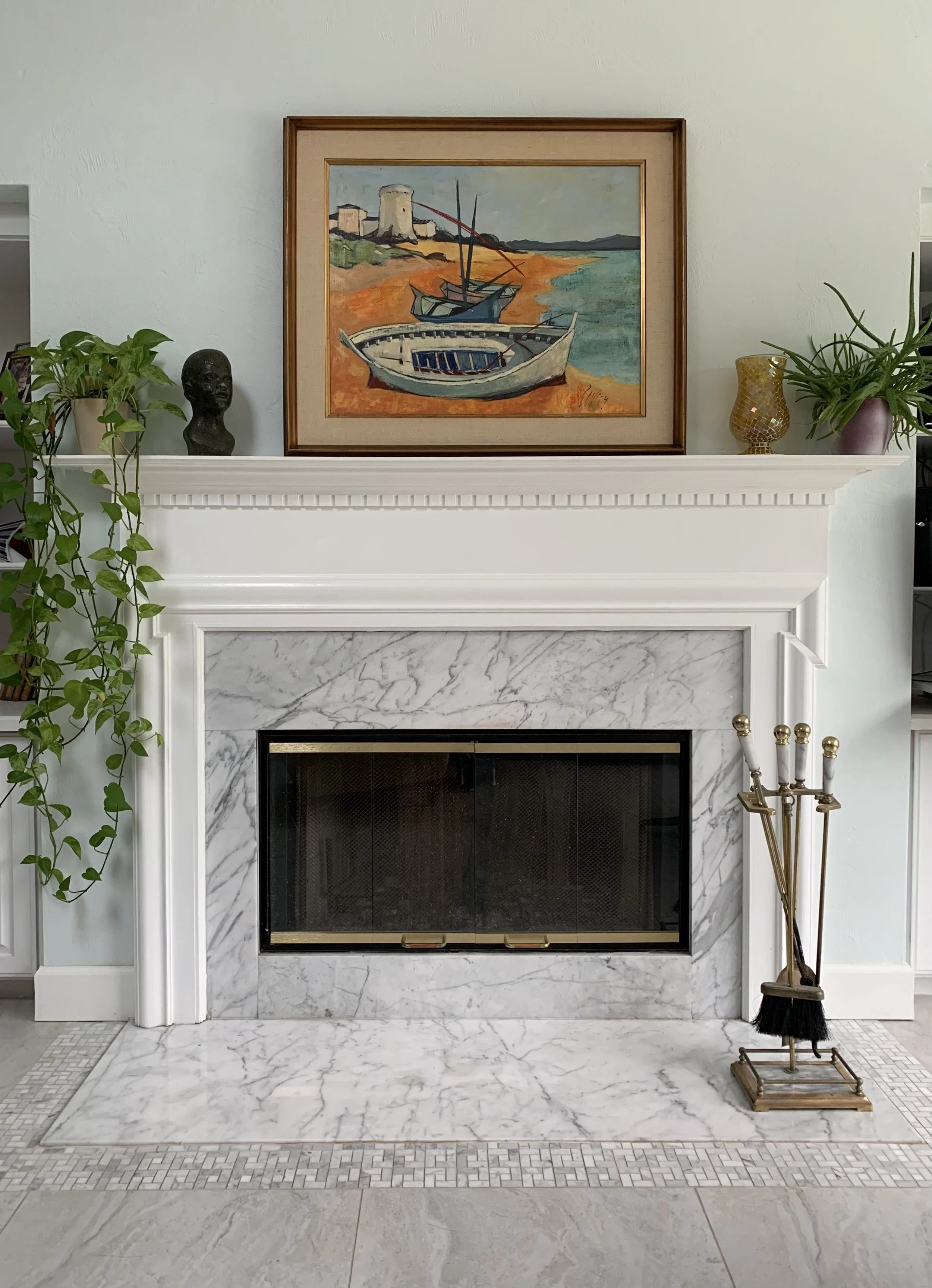 Living room fireplace with white marble surround, decorated with a framed vintage painting of boats on a French shoreline, potted plants, a small sculpture, and decorative vases, with fireplace tools on the right.