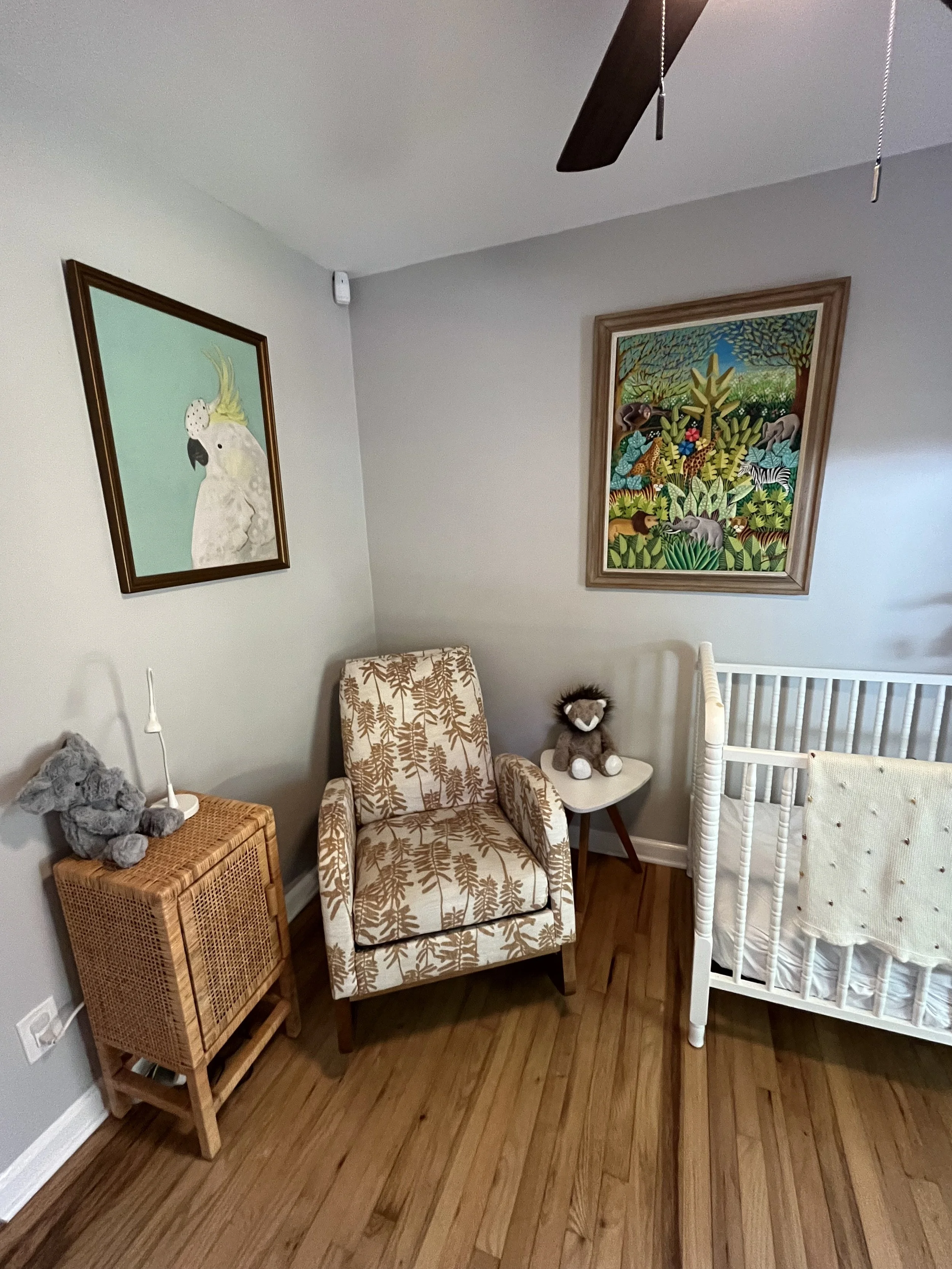 A cozy boho nursery corner with a patterned armchair, small side table with a stuffed animal, crib, colorful wall art, a ceiling fan, and a wooden floor.