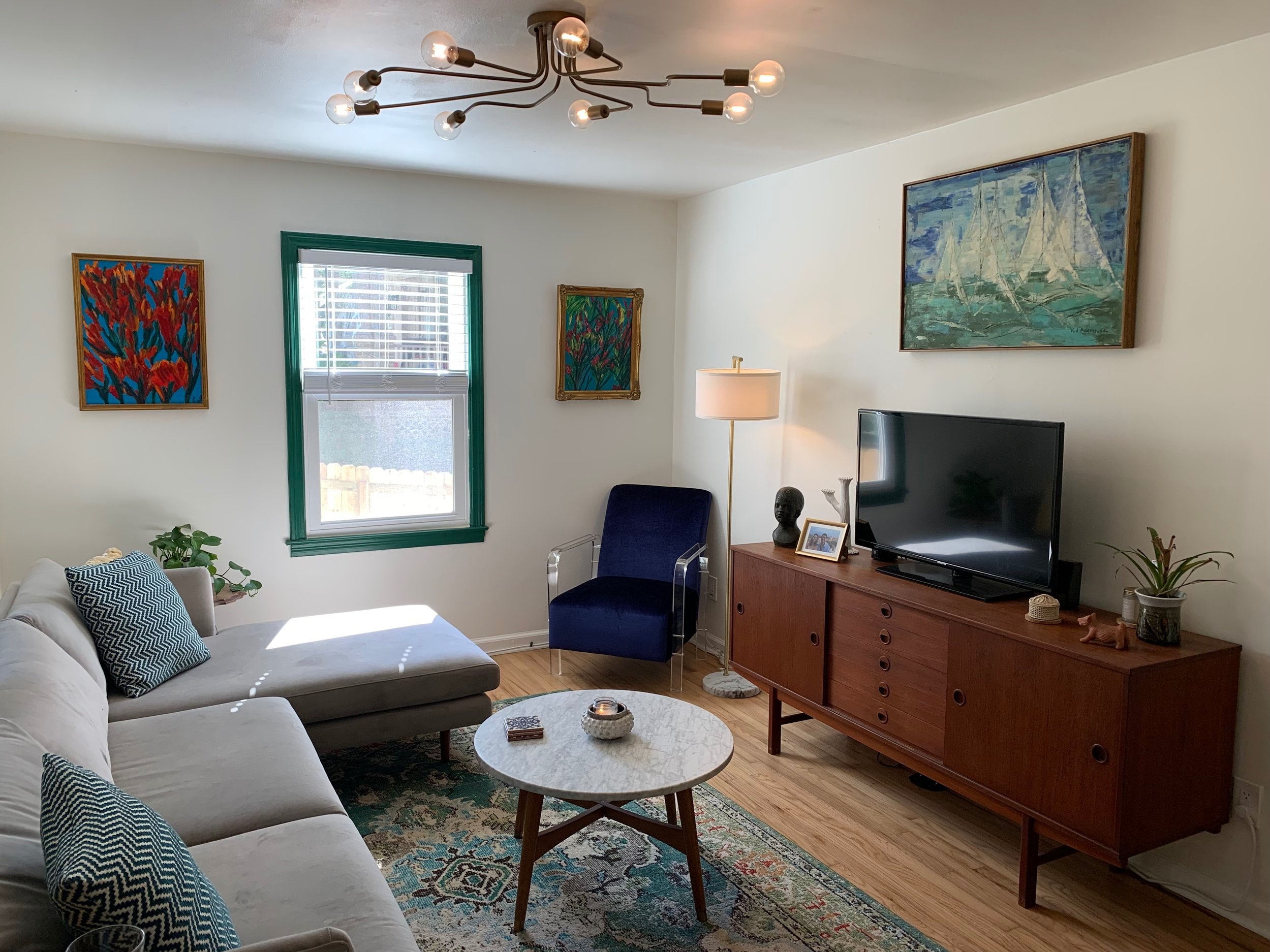 Living room with grey sofa, blue chair, wooden midcentury TV stand, flat-screen television, framed paintings, plants, rug, and a modern ceiling light fixture.