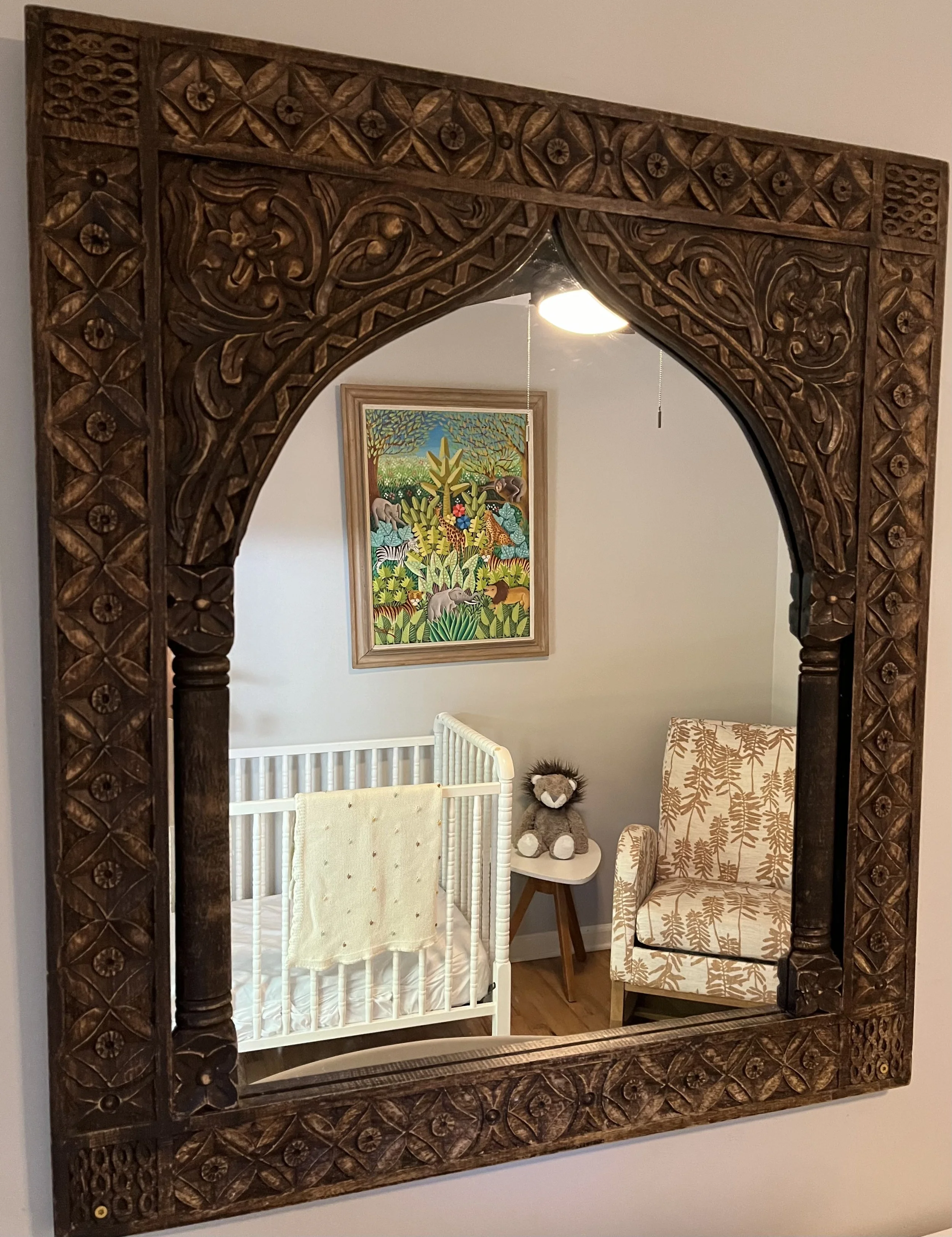 Moroccan mirror with intricate carved wood frame reflecting a nursery with a white crib, a plush lion stuffed animal, a tribal patterned armchair, a small rattan table, and a colorful jungle-themed painting on the wall.