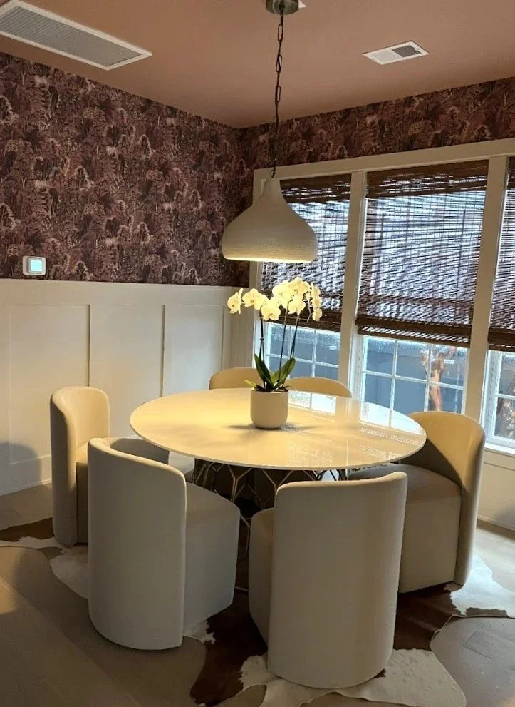 A bedroom with green walls and a bed with a beige headboard, gray bedding, white and patterned pillows, and two black nightstands with white lamps. There is a black and white abstract artwork on the wall above the bed, a ceiling fan, and windows with white curtains.