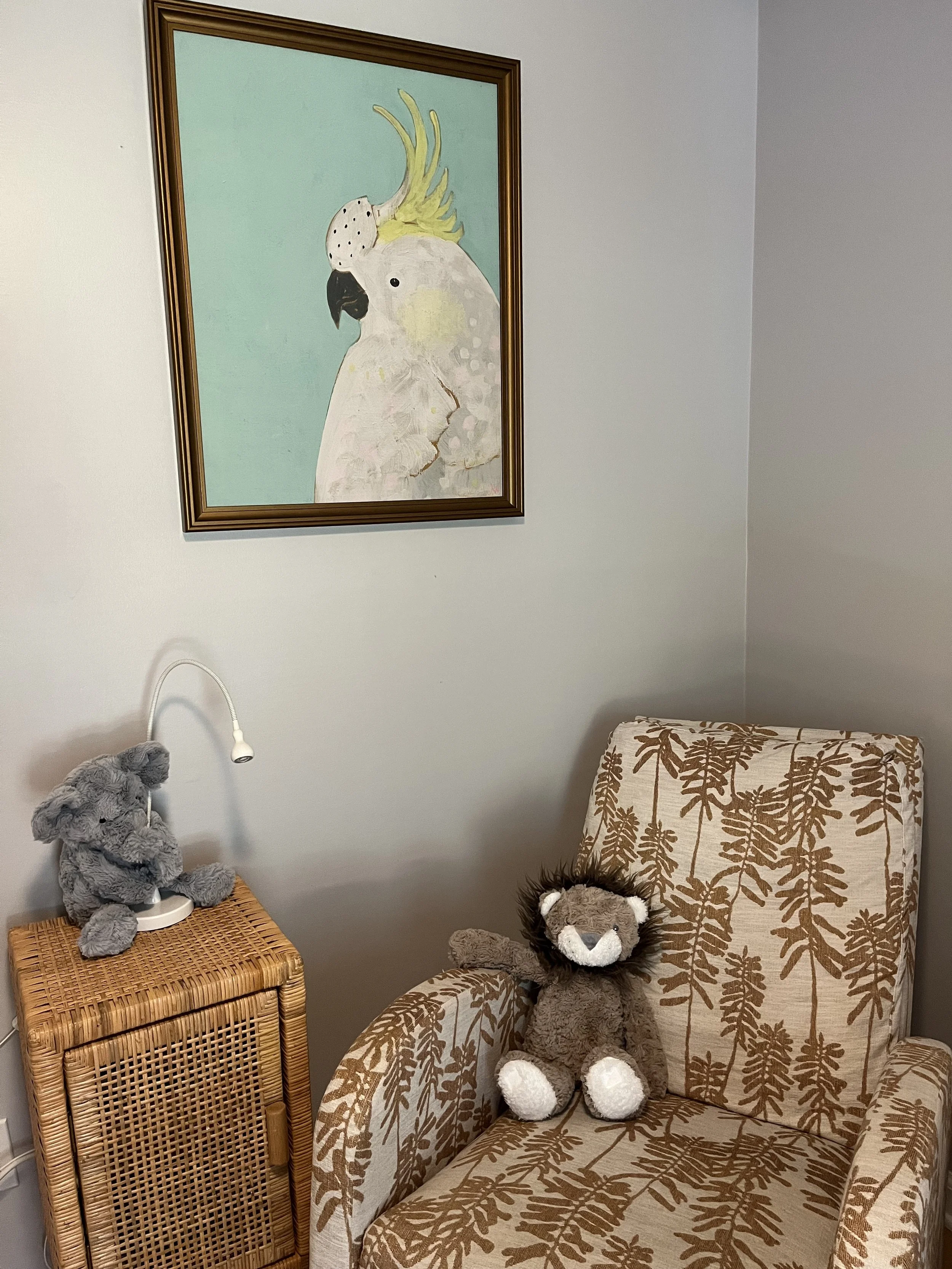 A cozy corner with a patterned armchair, a stuffed lion toy, a small ruffled stuffed bear on a wicker side table with a lamp, and a framed painting of a white cockatoo on a light green background hanging on the wall.