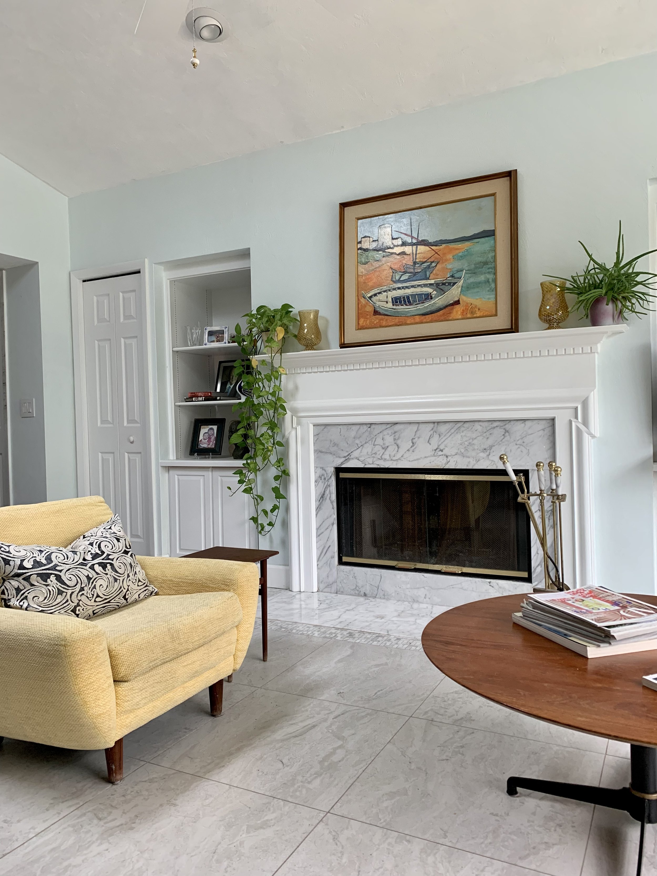 Living room with midcentury yellow armchair, fireplace with marble surround, framed painting of boats on water, plants and decorative items on mantel, shelf with picture frames and books, and a wooden midcentury coffee table with magazines.