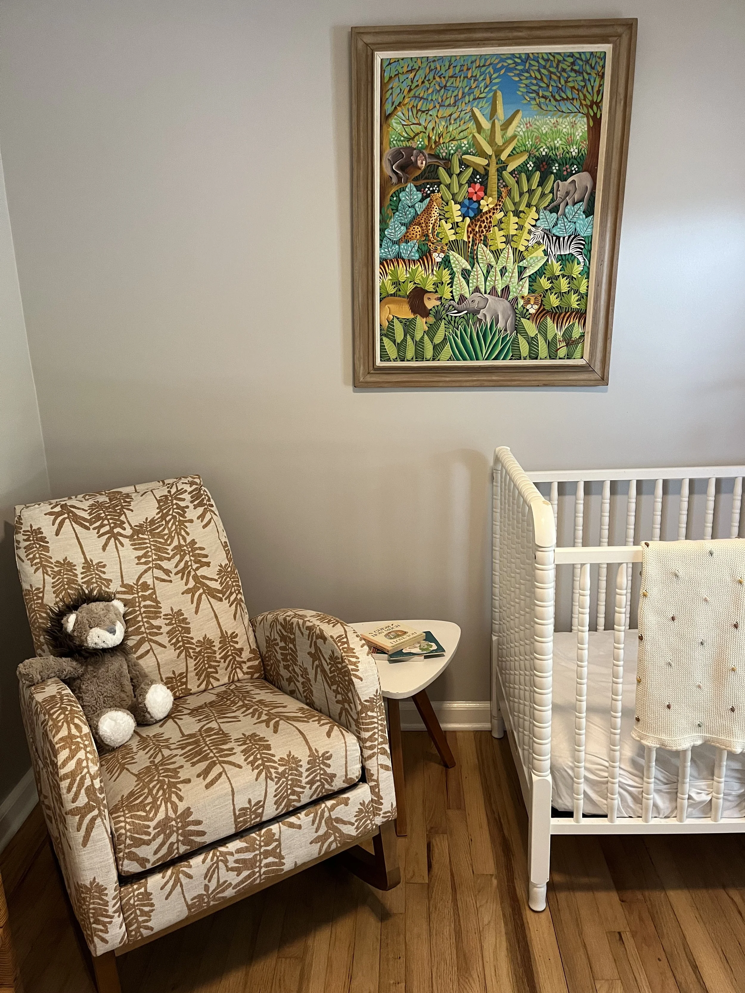 A nursery with a patterned armchair, a small white side table, a crib, and a colorful jungle-themed painting on the wall.
