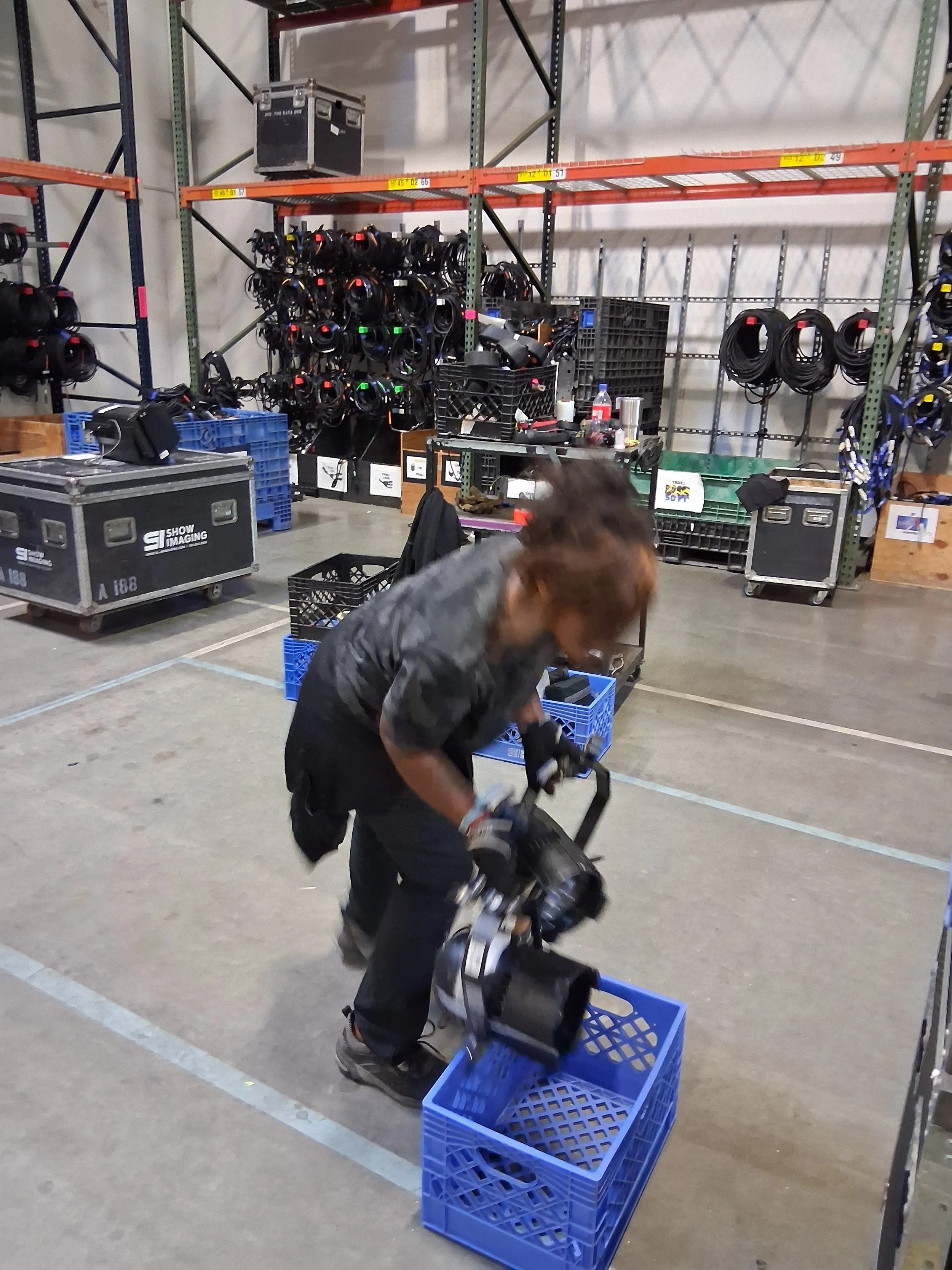 Warehouse worker organizing equipment, loading cases, and managing inventory in a production warehouse with lighting gear and staging materials.