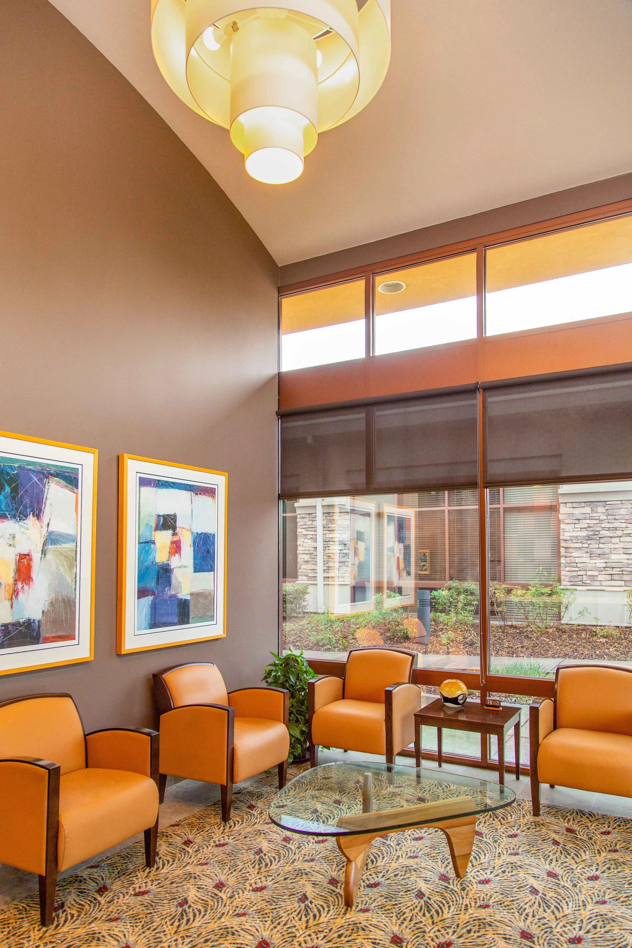 A waiting area with yellow leather chairs arranged around a glass coffee table. The wall has two colorful abstract paintings framed in yellow. Large windows show an outdoor brick building and landscaped area with plants. A small table with a decorative object and a plant is also visible.