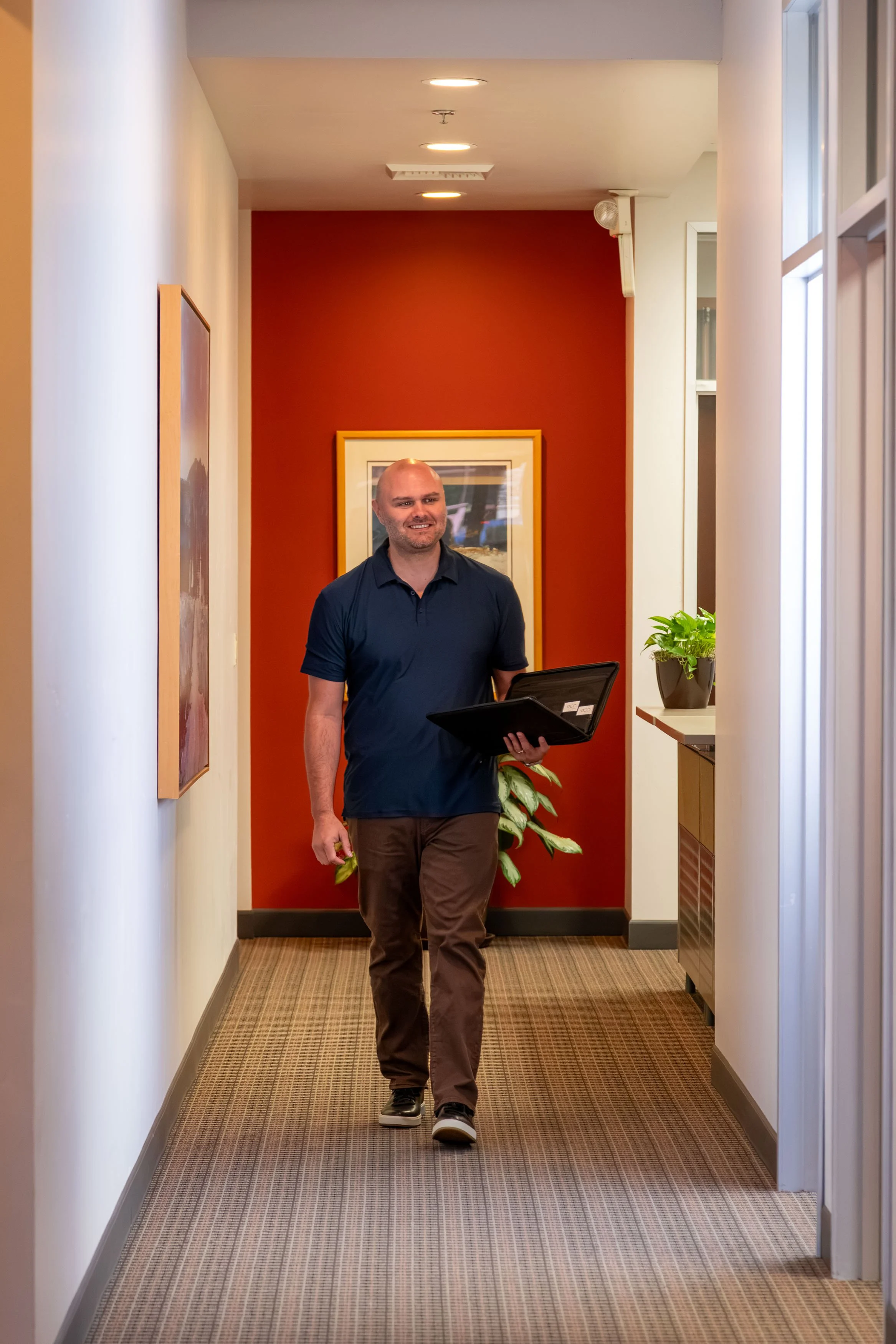 A man in a dark blue polo shirt and brown pants is walking down a hallway while holding a black briefcase in his right hand. The hallway has a beige carpet, white walls, and a red accent wall at the end with framed artwork. There is a potted plant on a small table on the right side.