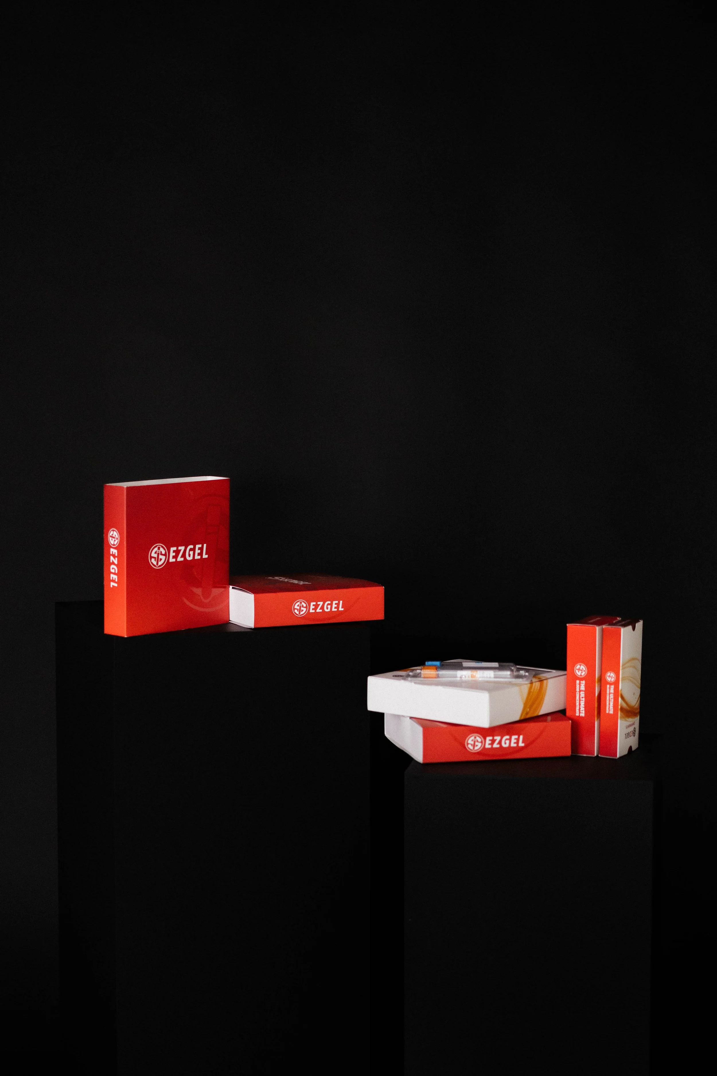 Boxes of EZGEL products and syringes displayed on black pedestals against a black background.
