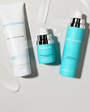 Skincare products including Neo Cleanse gentle cleanser, Lumior firm eye cream, and Bio Cream firming and brightening cream on a white surface with some cream spill.