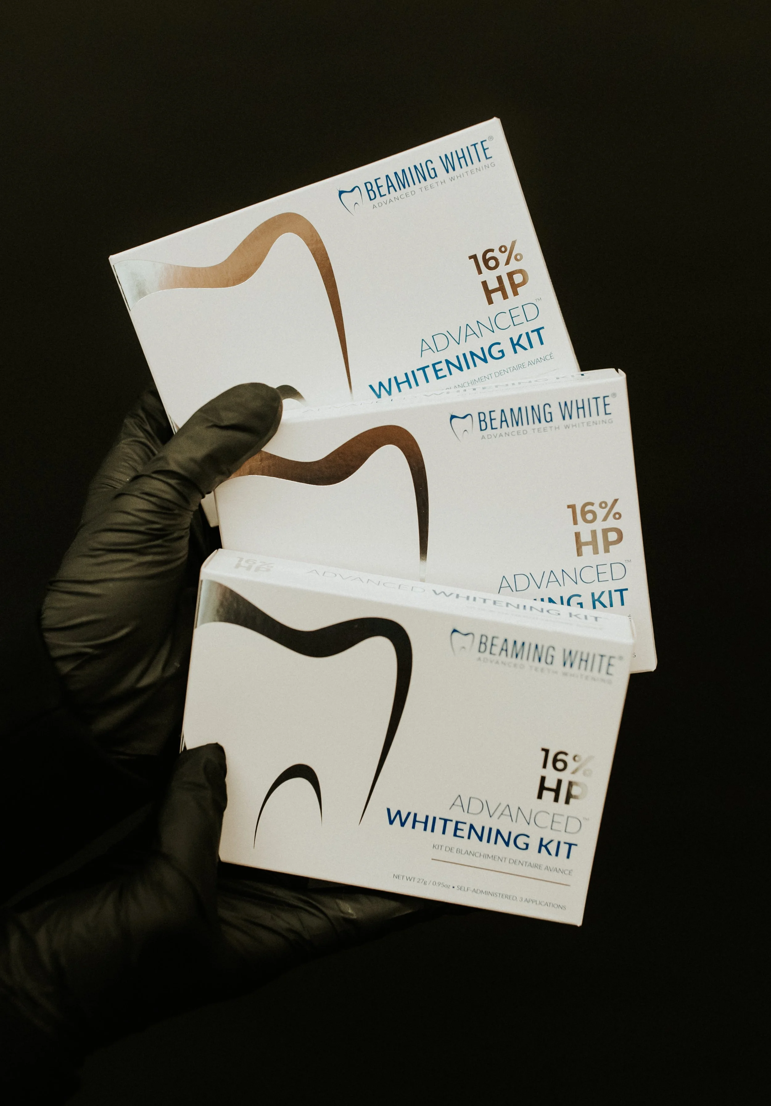 A person wearing black gloves holding three boxes of Beaming White advanced teeth whitening kits against a black background.