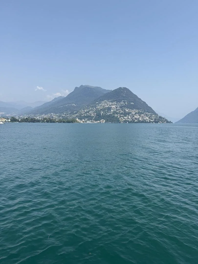 This is broad view of Lake Brienz, Switzerland, taken by Dr. Namita Sethi.  The focus of the picture is a cluster of mostly green hills, with homes at their bases.  One also sees blue sky and a large calm water area in the foreground.
