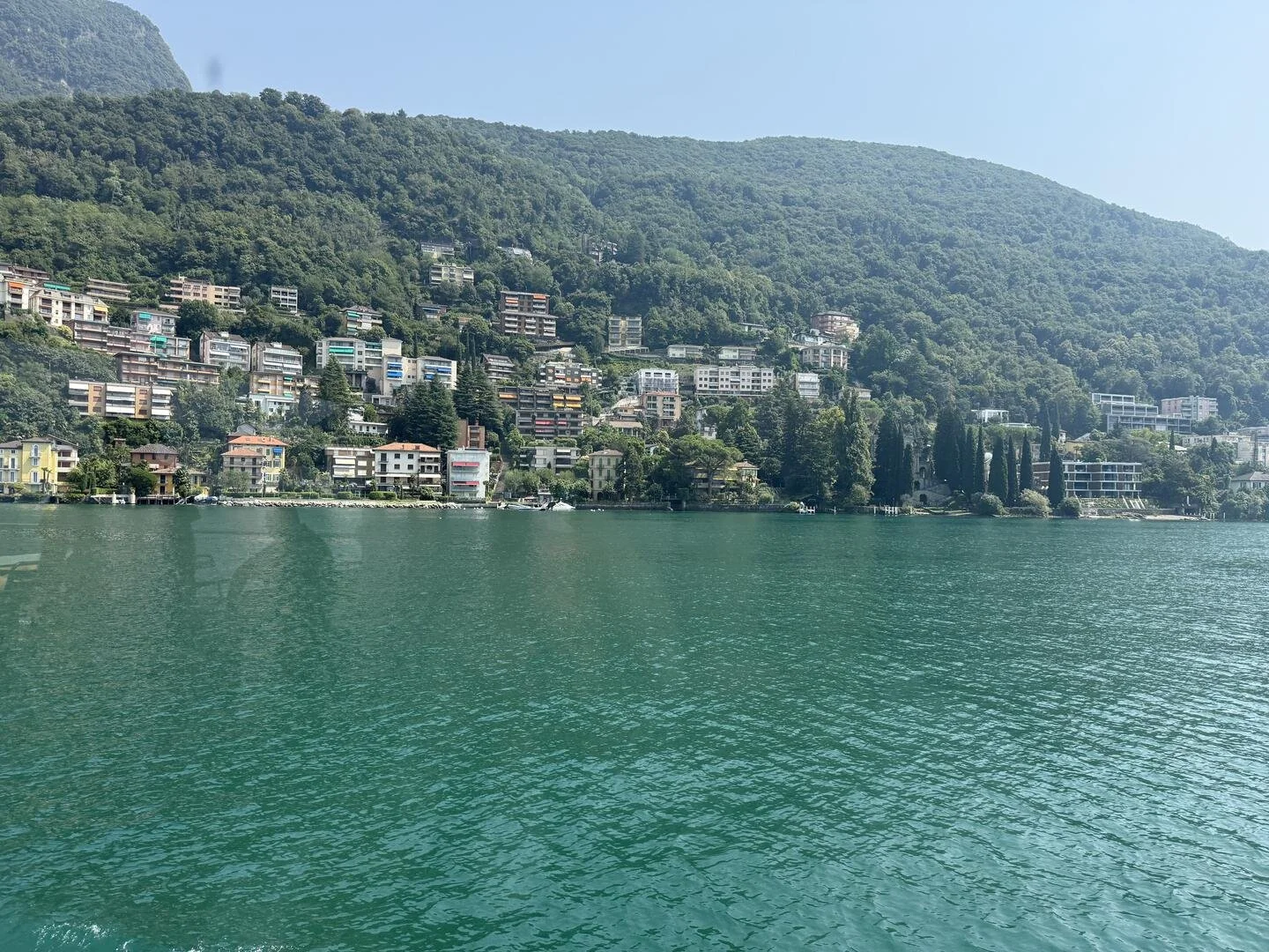 This is broad photo of Lake Brienz, Switzerland, taken by Dr. Namita Sethi.  The photograph shows a green mountain and hill-base houses in the background; in the foreground one sees greenish water.  One can also see a part of the blue sky.