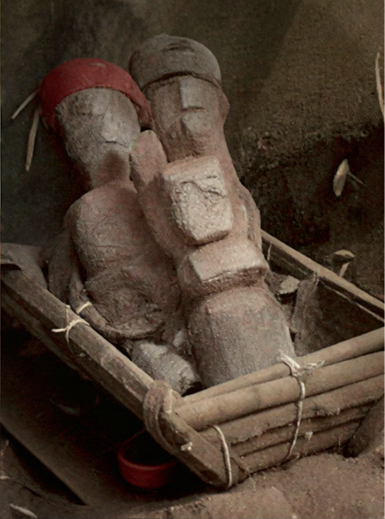 Three wooden doll-like figures that are part of a traditional, pre-religious ritual called Agwu; they appear to represent mother, father, and child, with the mother figure sporting a red head gear; they are all placed in a wooden basket in this image