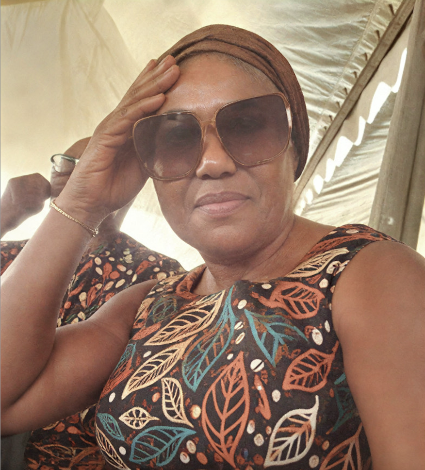 This is a Nigerian woman with a headgear and a patterned, colourful sleeveless upper-body garment; she is sporting big sun glasses and smiling.  Her name is Vivian Ottih.
