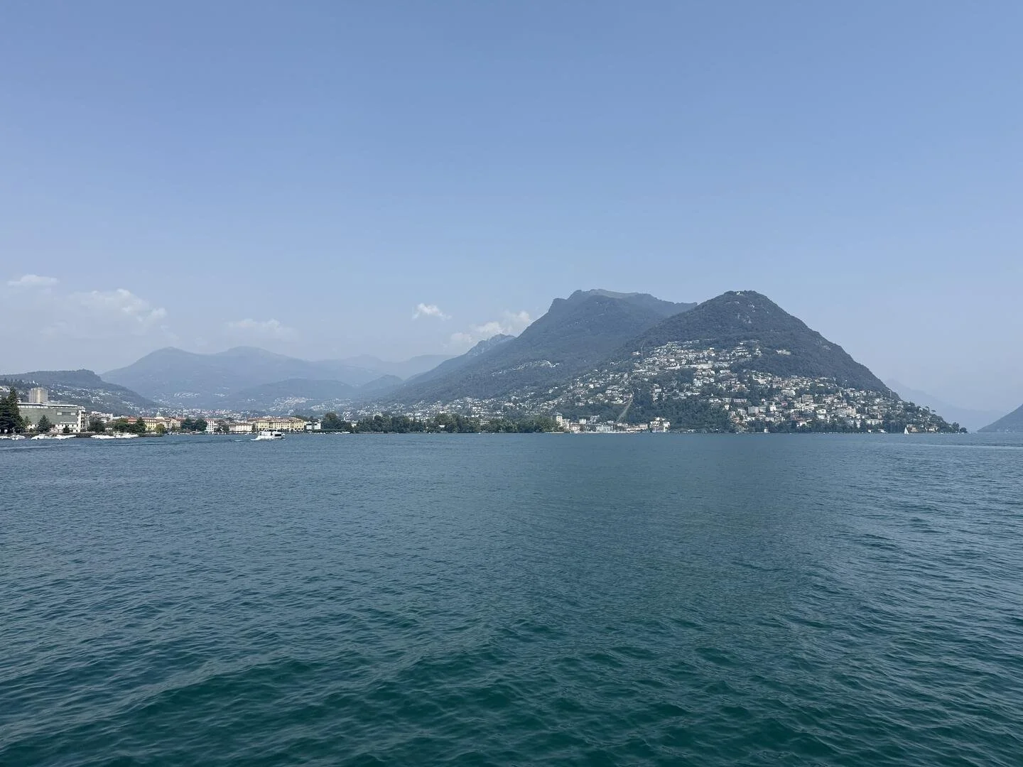 This is broad view of Lake Brienz, Switzerland, taken by Dr. Namita Sethi.  The photograph shows bluish hills, clouds, a blue sky, many hill-base homes in the background; in the foreground one sees calm bluish water.