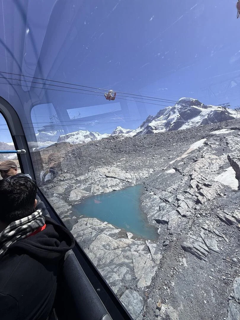 Taken from inside an aerial gondola, this image shows a part of the ropeway infrastructure in Zermatt, Switzerland, underneath a blue sky.  One can also see a rocky area, a pool of water, and snow-covered hills.  Two human males are also seen inside.