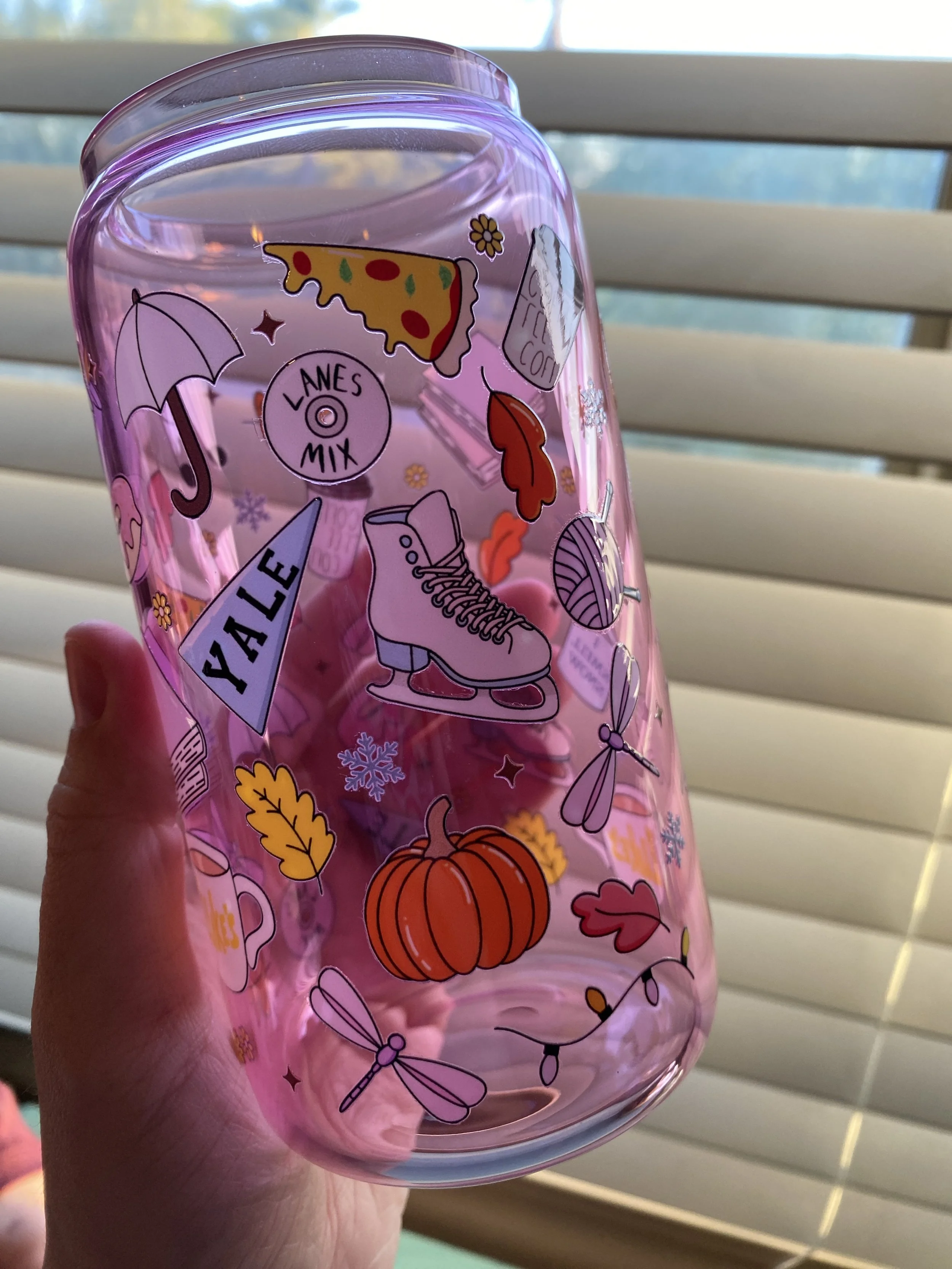 Pink glass with fall-themed stickers including pumpkin, leaves, ice skate, umbrella, pizza, snowflake, and a djinn, held in front of white window blinds.