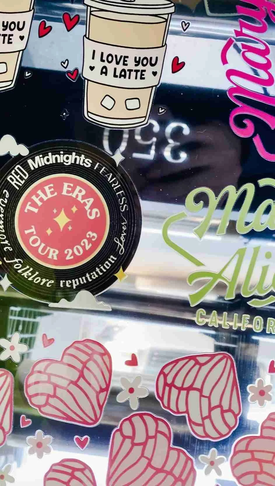 Decorative stickers on a glass surface, including a coffee cup with the text 'I love you a latte,' a circular emblem for The Eras Tour 2023, and pink heart-shaped designs with white stripes.