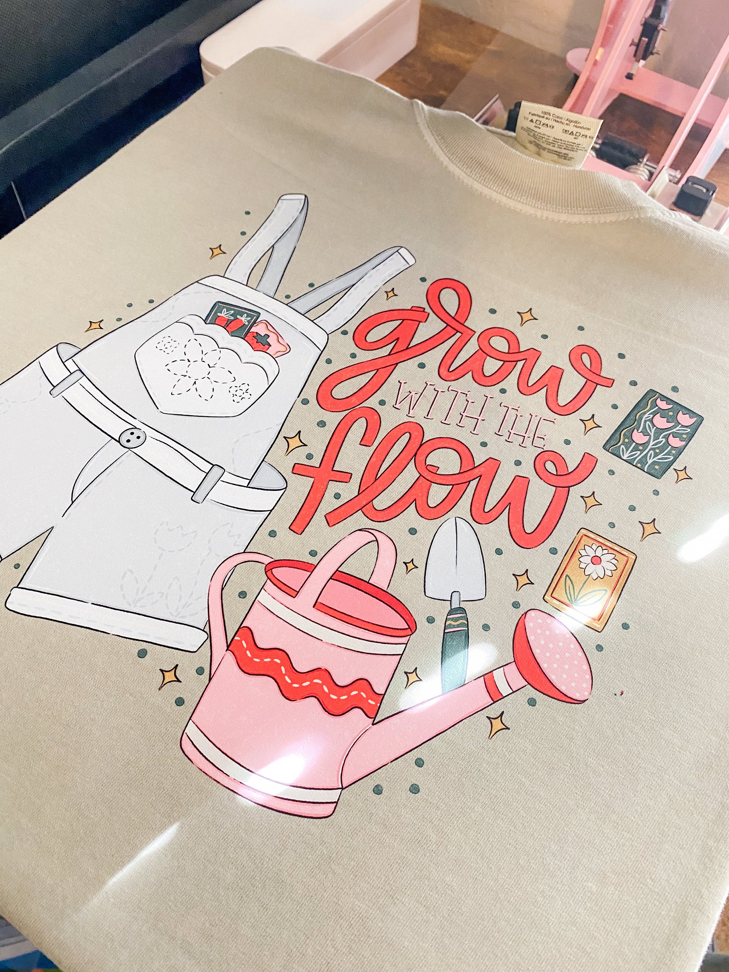 Beige sweatshirt with gardening-themed design, including overalls, watering can, trowel, seed packet, and flowers. Text says 'Grow with the Flow.'