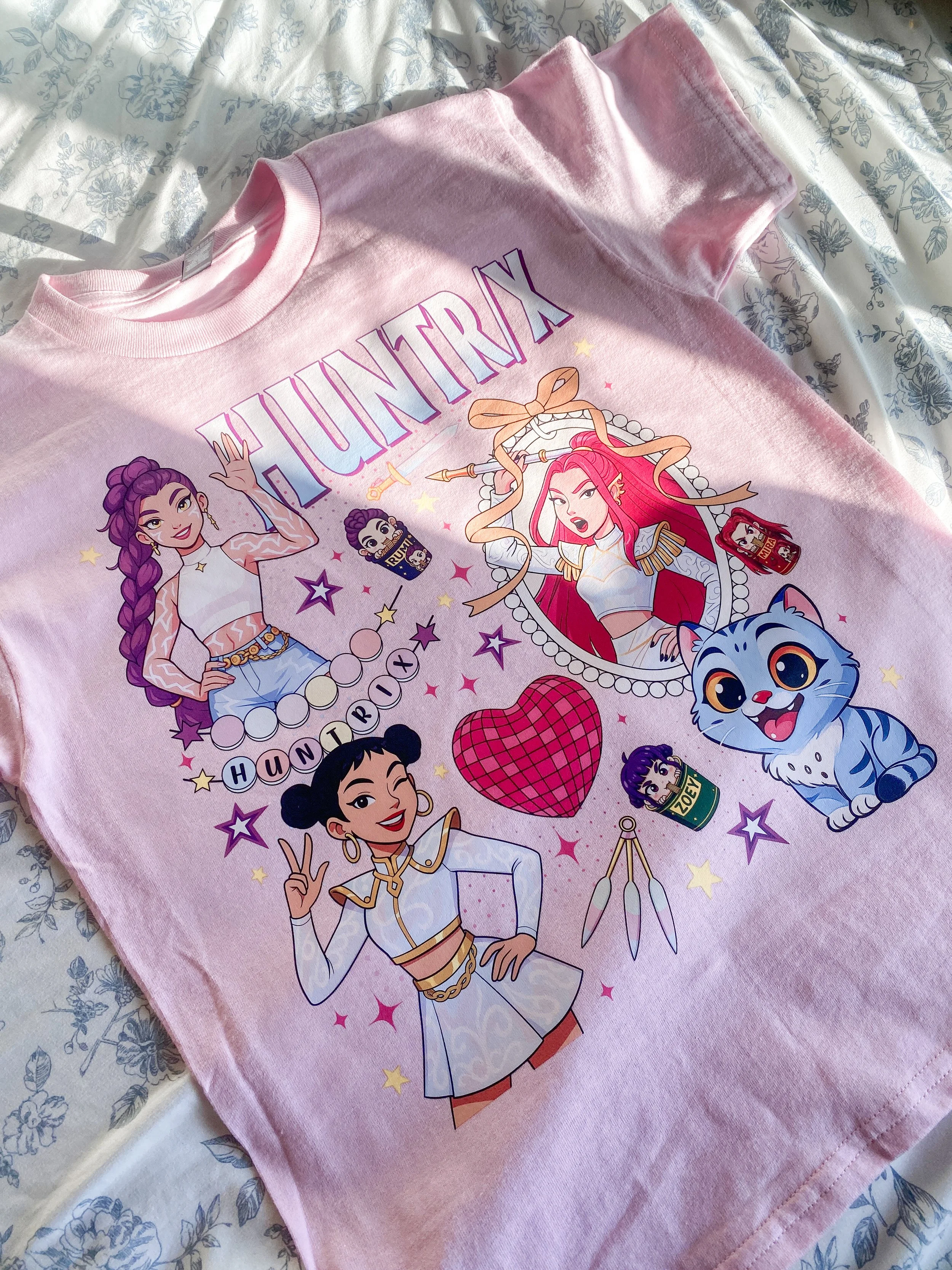 Pink T-shirt with animated characters from the show "Huntix" and various symbols, stars, and a heart printed on it.