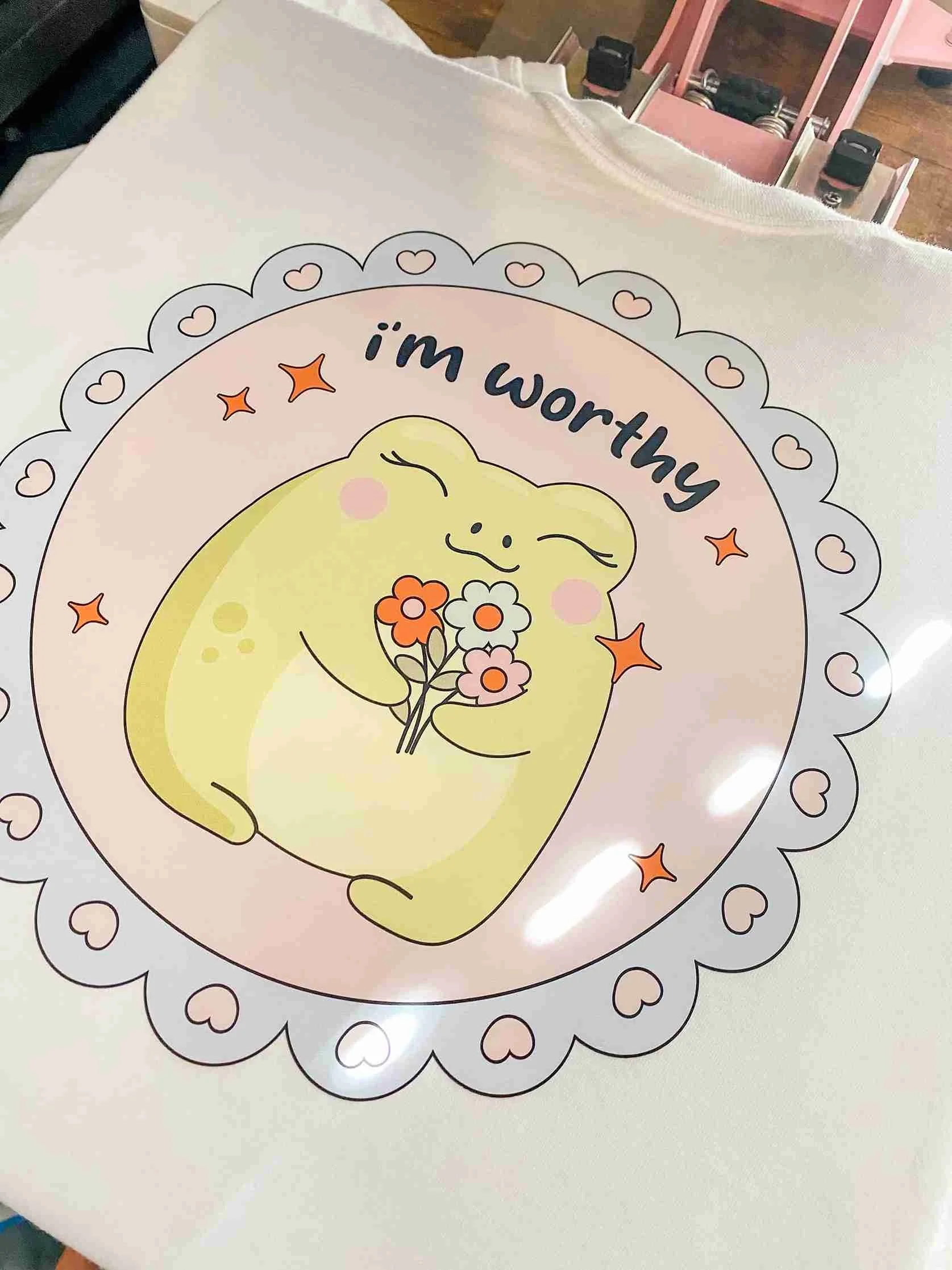 A circular sticker featuring a cute, smiling yellow frog holding a bouquet of pink and orange flowers, with the words 'I'm worthy' above it and a border of small hearts and orange stars.