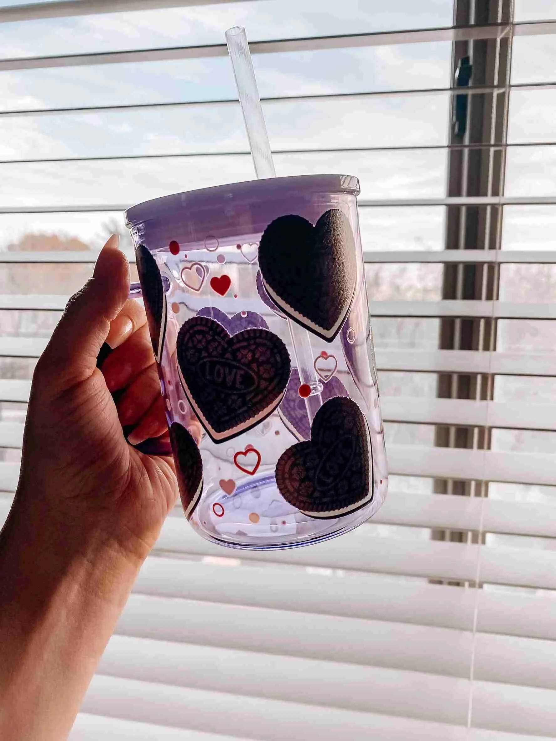 A transparent tumbler cup decorated with black heart-shaped cookie stickers, and smaller red and pink hearts, held by a hand in front of window blinds with an overcast sky in the background.
