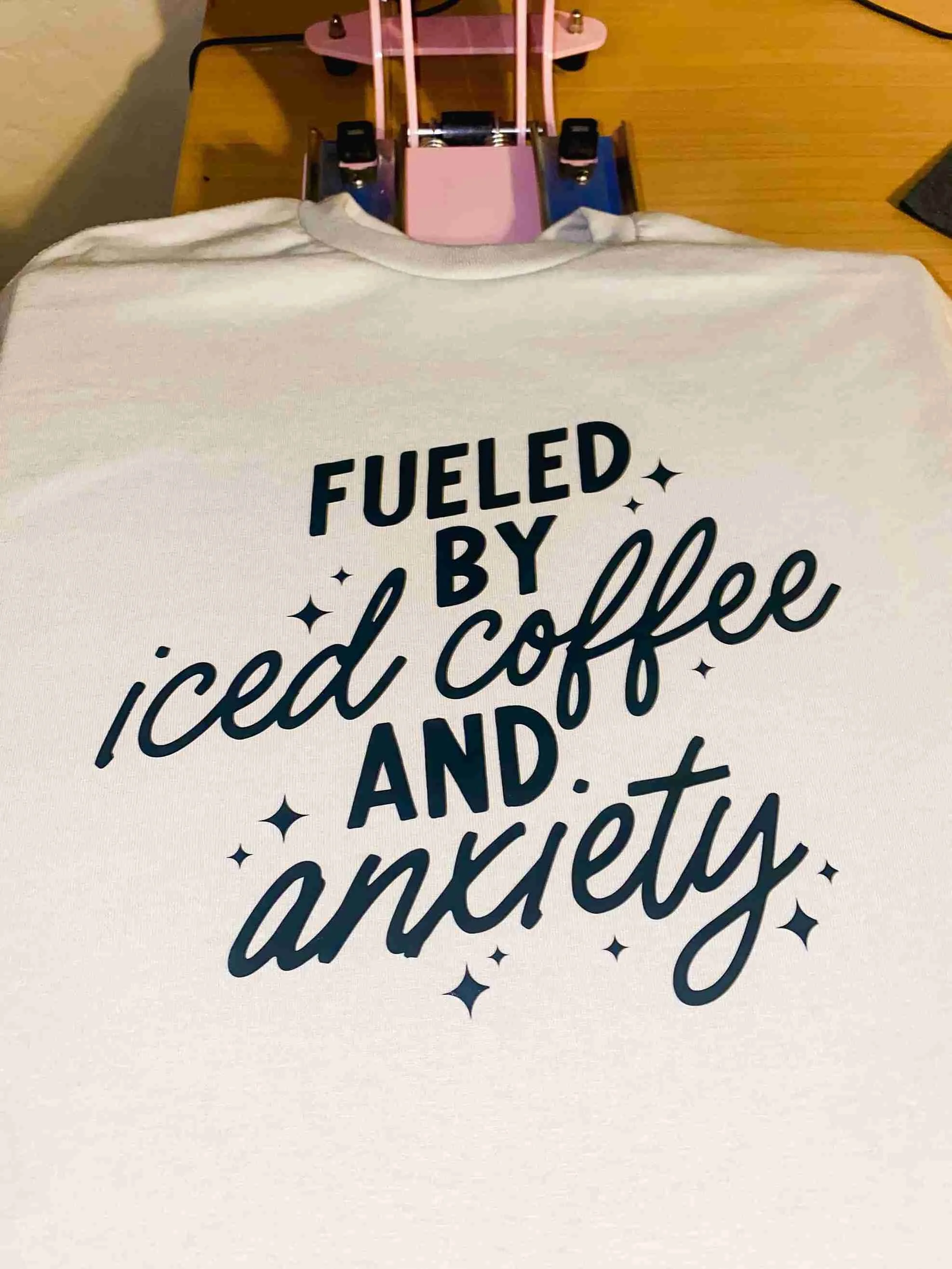 White t-shirt with the text "Fueled by iced coffee and anxiety" in black lettering, placed on a wooden table with a pink and black hot glue gun in the background.