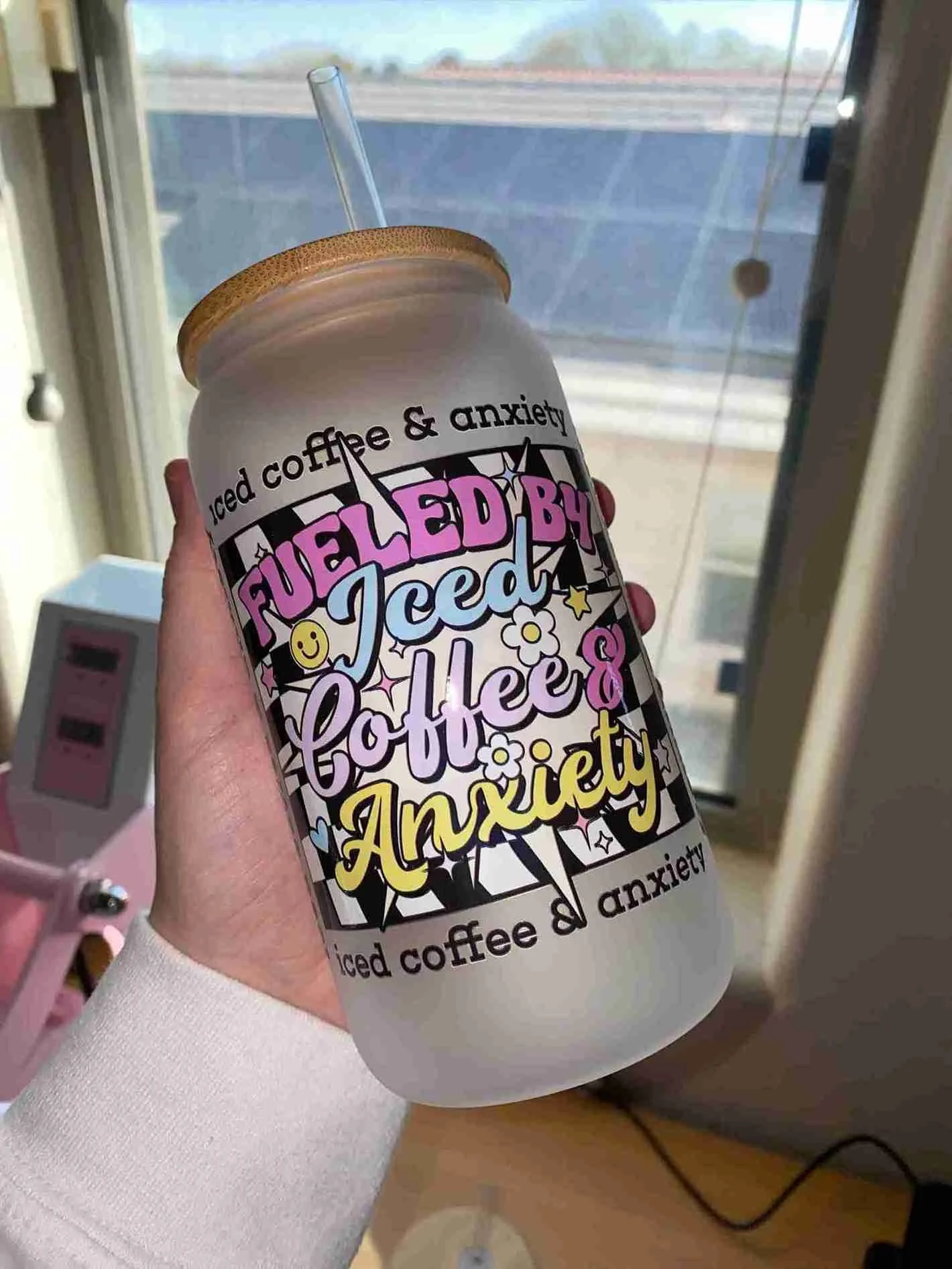 A hand holding a white tumbler with a colorful, cartoon-style design that reads, "Fueled by iced coffee & anxiety." The tumbler has a clear straw and a wooden lid, with a window and a cloudy sky in the background.
