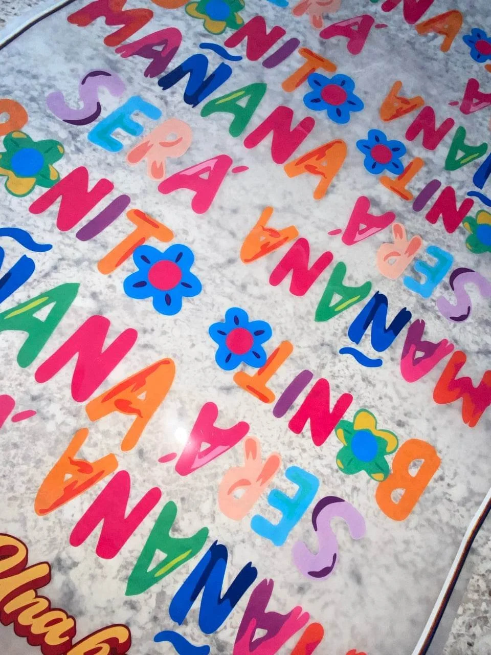 Colorful paper cutouts with the repeated word 'MANA' and various flower illustrations on a silver background, likely part of a festive decoration.